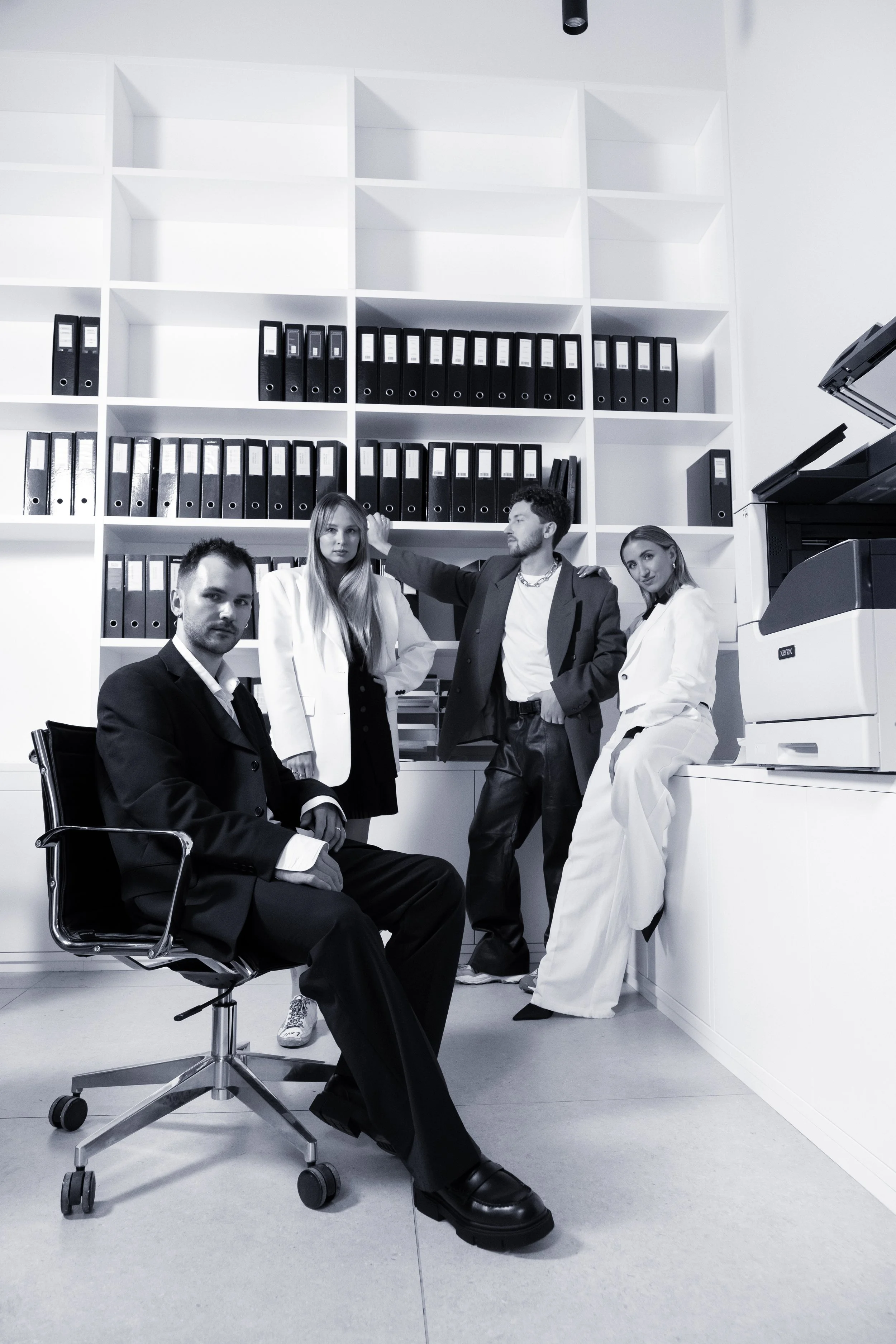 Four young professionals dressed in formal, stylish clothing in an office environment with a white wall of shelves filled with binders, a desk, and a high-tech printer.