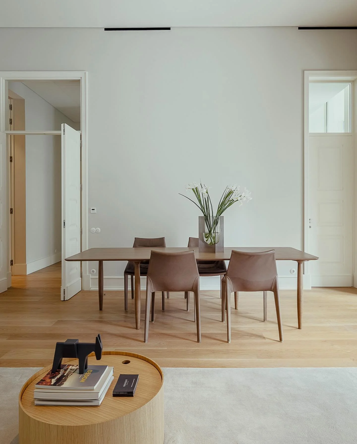 Minimalist dining room with a wooden table, four brown chairs, a large vase with white flowers, and a light-colored rug on wooden flooring.