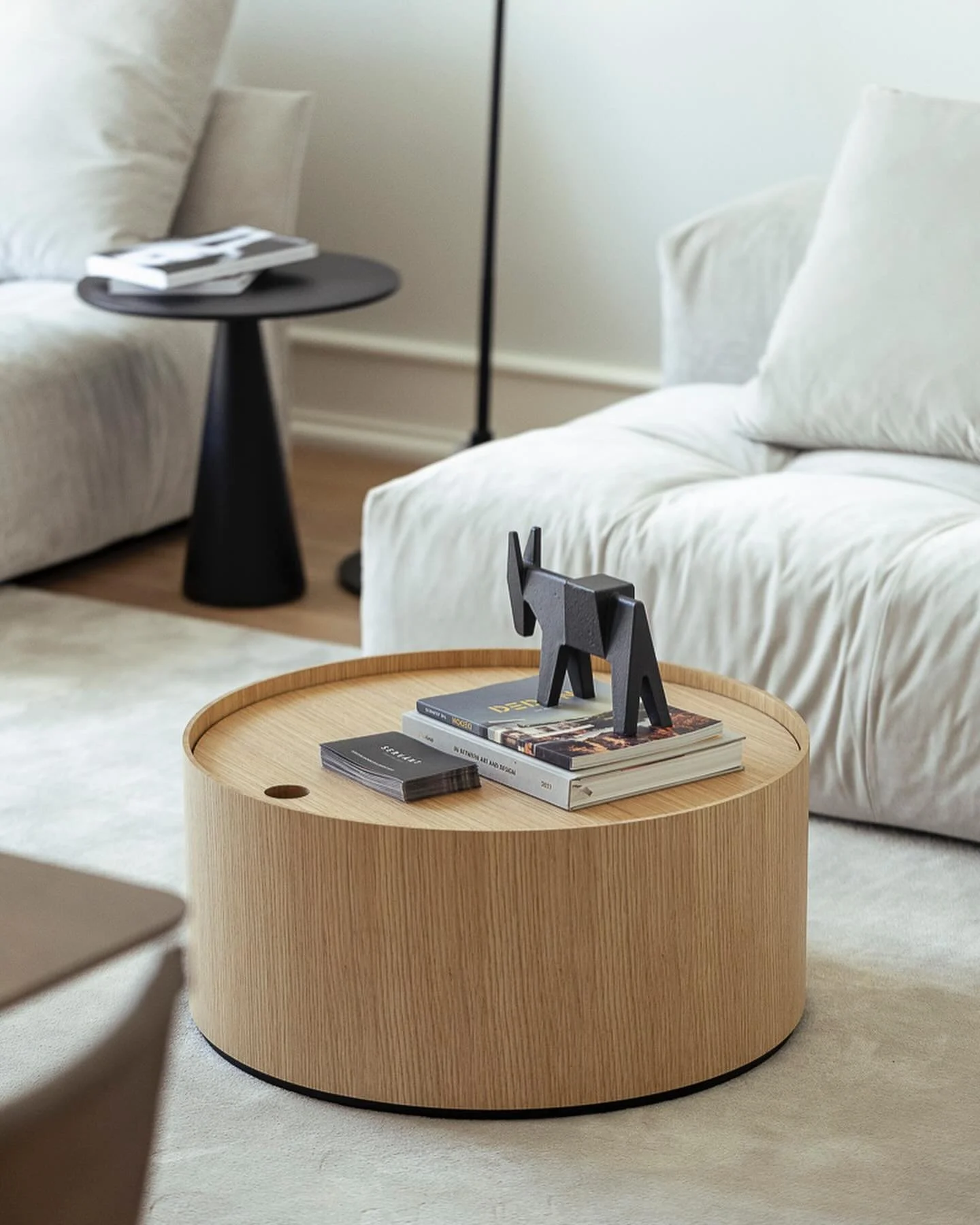 Wooden round coffee table with stacked magazines and a black decorative elephant figurine in a modern living room with white sofas and black side table.
