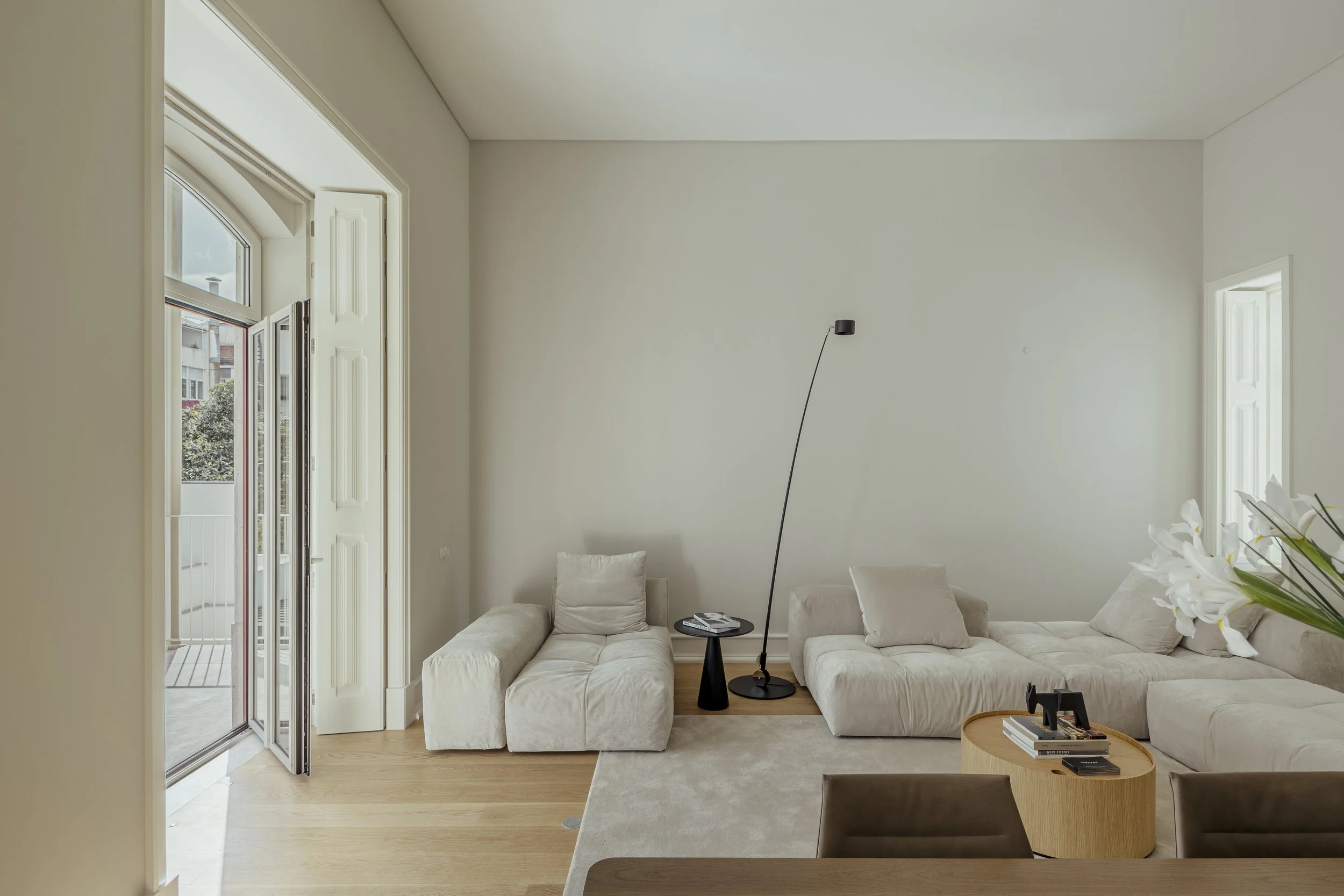 Minimalist living room with white sofas, a small black side table, a tall black floor lamp, a round wooden coffee table, and a vase with white flowers. Large windows with open shutters let in natural light.