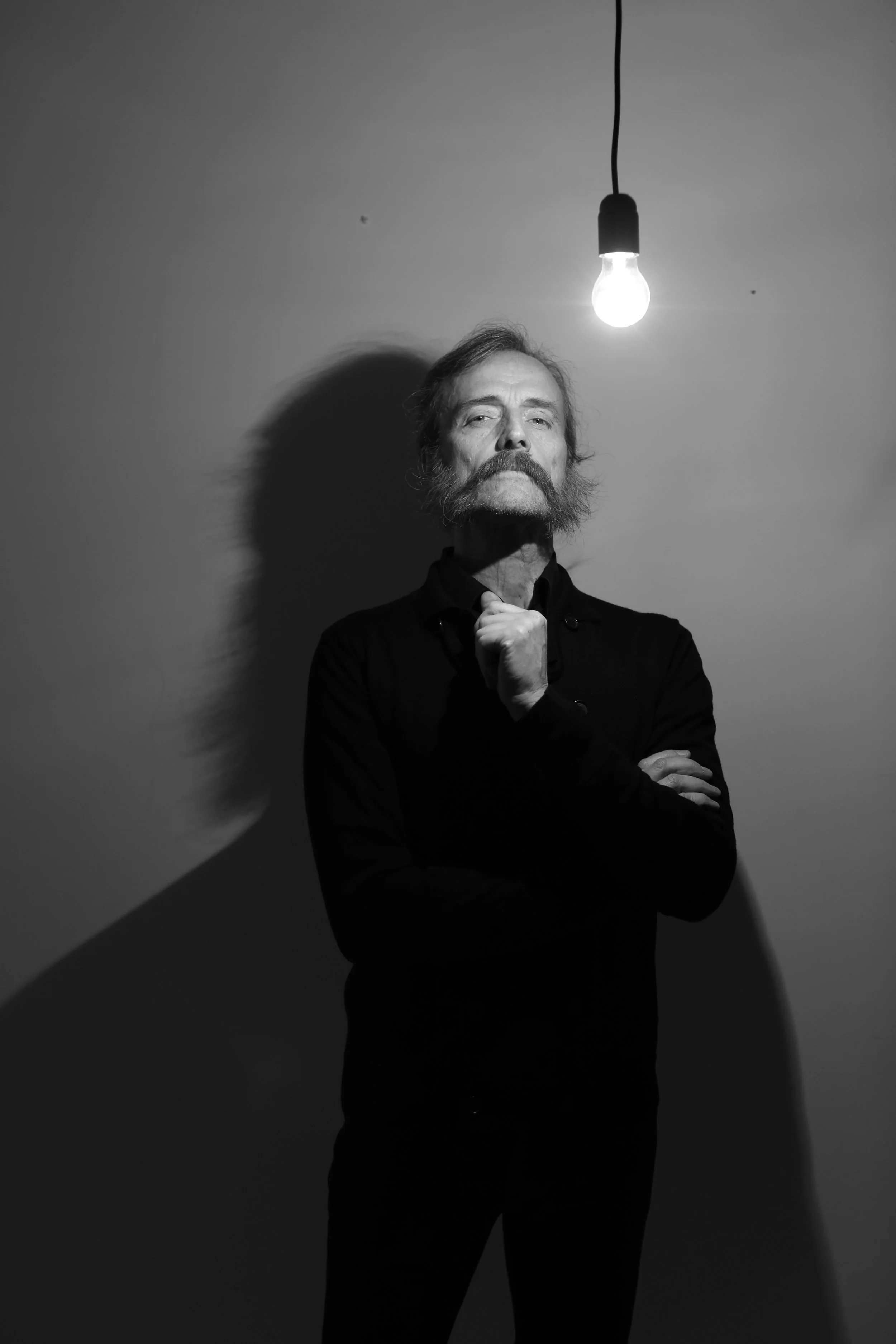 Black and white photo of a man with a beard and mustache standing under a lit light bulb hanging from the ceiling, with one hand on his chin and the other arm crossed.
