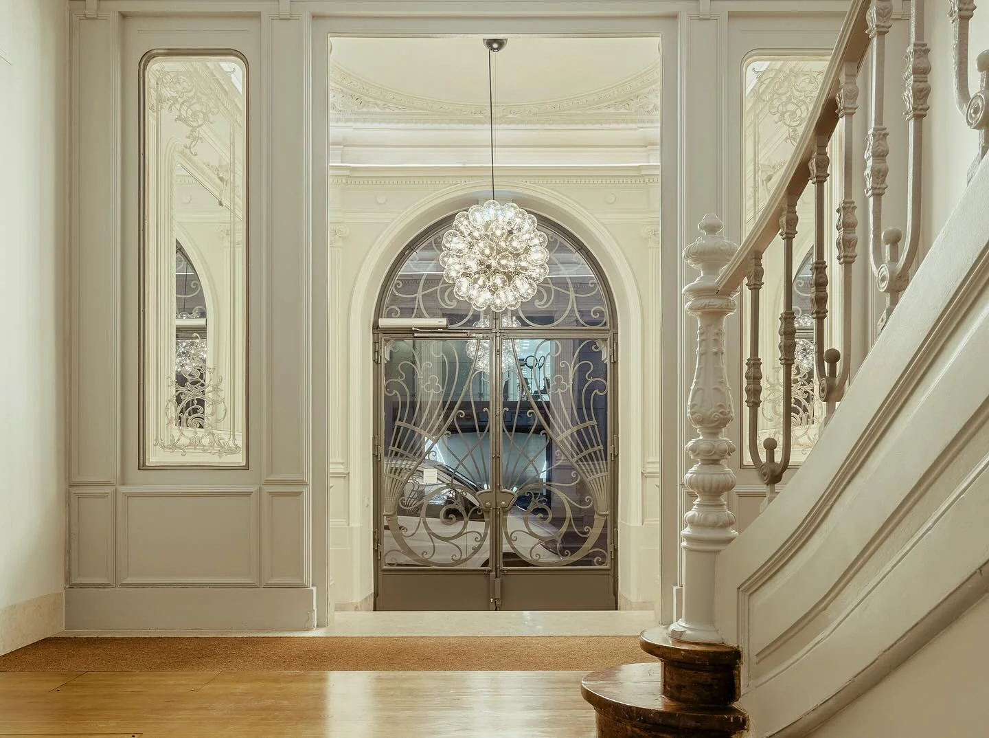 Elegant entryway with ornate white walls, glass-paneled double doors with intricate metalwork, a crystal chandelier, and a curved staircase with a white banister.