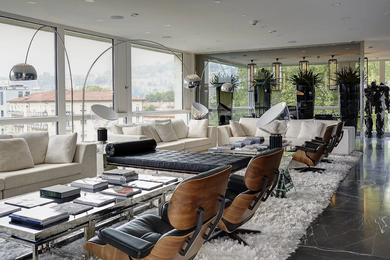 Modern living room with large windows, white sofas, wooden chairs, black coffee table, and decorative lighting fixtures.