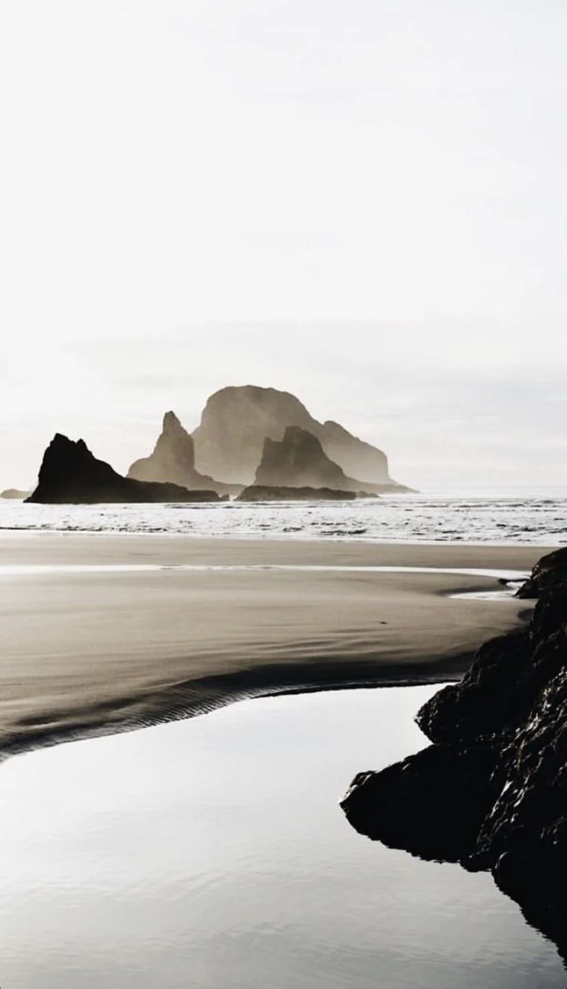 A black and white photograph of a beach with large rock formations emerging from the water in the background, and a sandy shoreline with rocks and a calm reflective pool in the foreground.