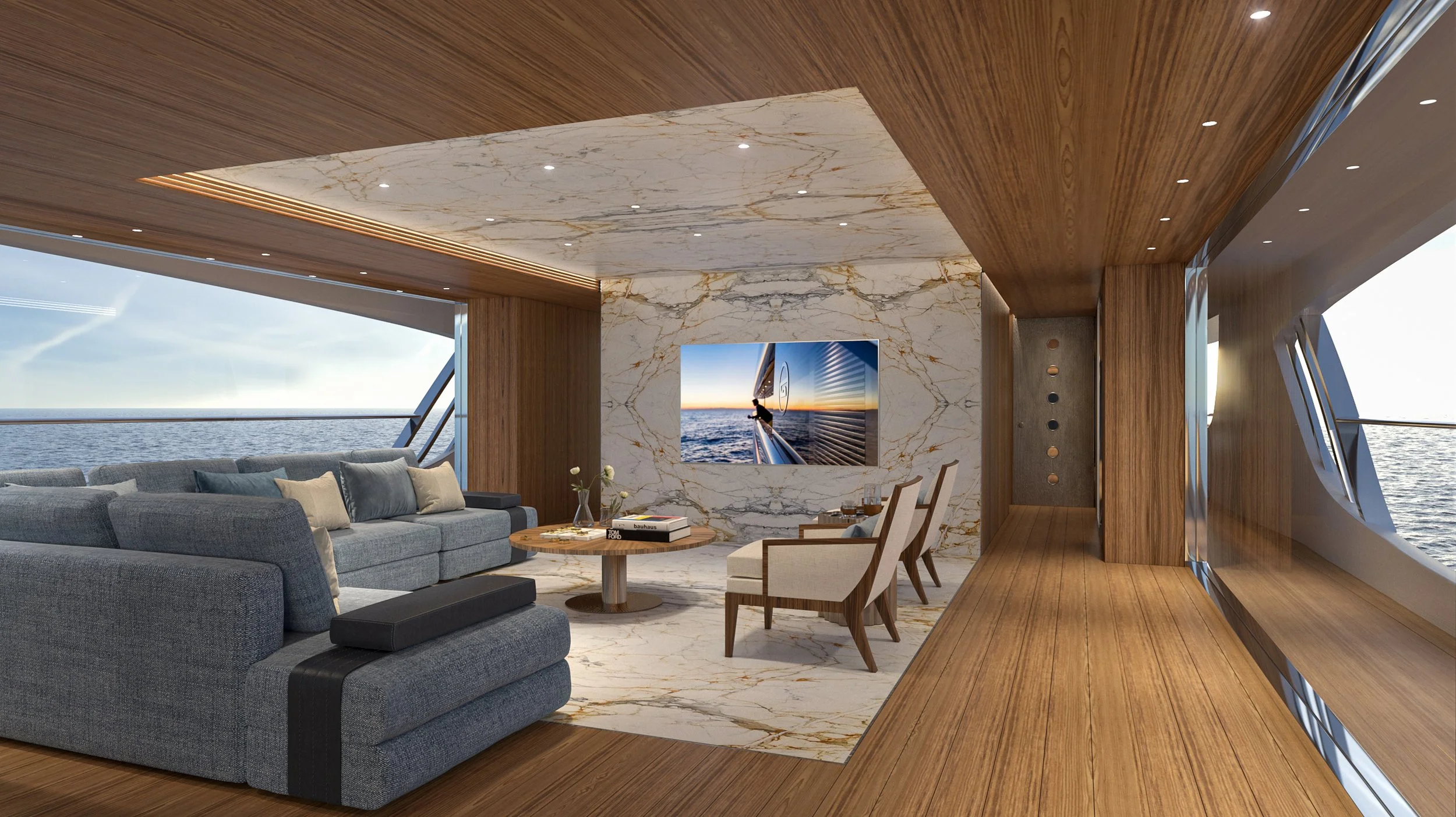 Inside a yacht with a modern living area, featuring a large gray sofa with cushions, a round wooden coffee table with books and a vase of white flowers, two chairs with beige upholstery, a marble accent wall with a large flat-screen TV, and panoramic windows showing the ocean.
