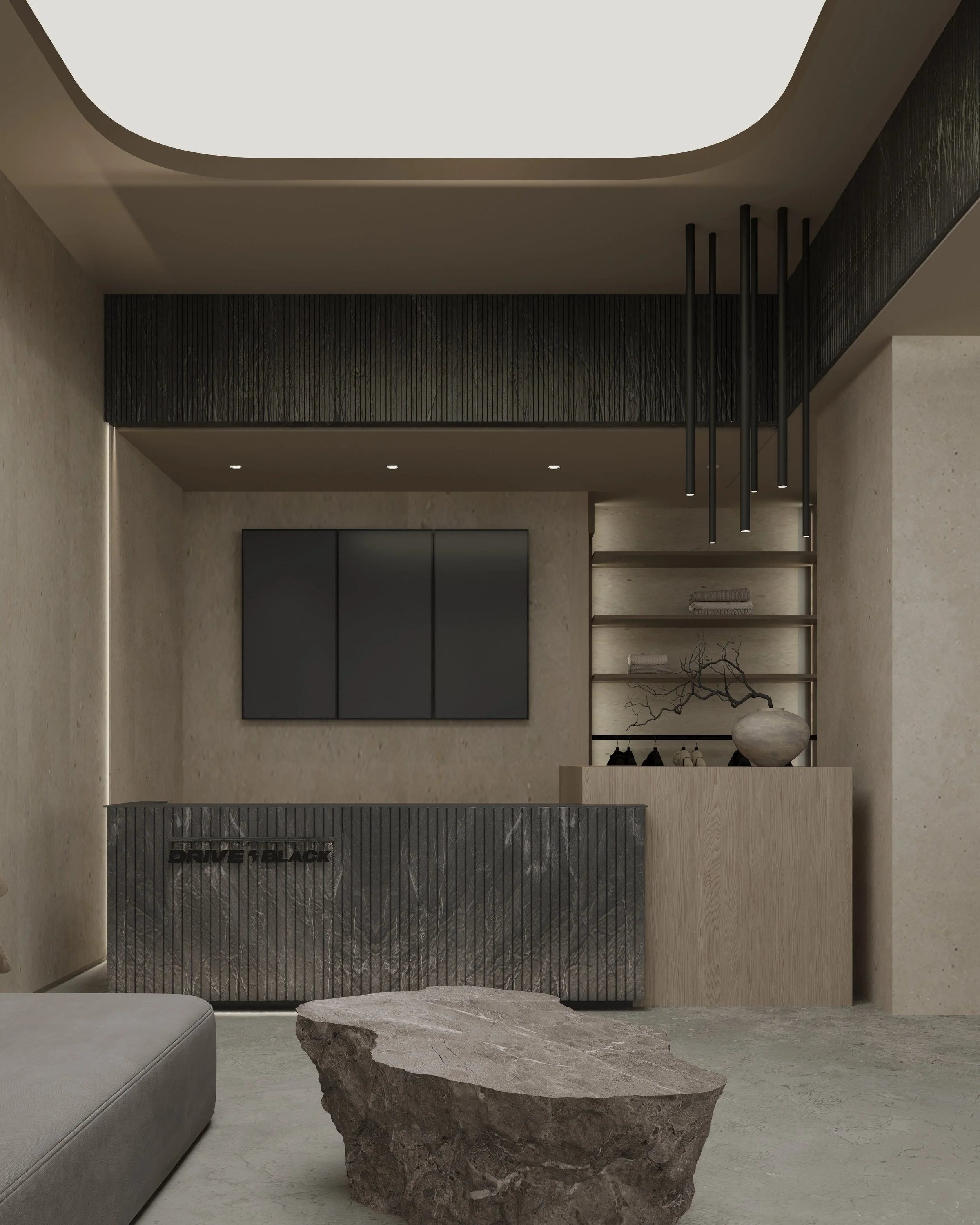 Modern interior space featuring a textured gray stone reception desk with the phrase 'DRIVE BLACK' on it, a large rough-textured stone on the floor, black shelving with decorative objects, a minimalist wall-mounted TV, and black pendant lights hanging from a ceiling with trim lighting.