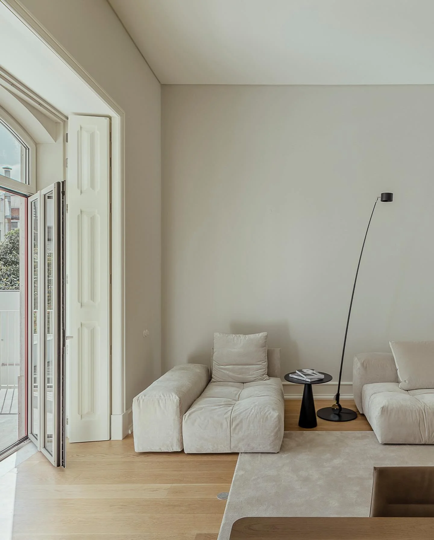 A corner of a living room with two cream-colored sofas, a small black side table with magazines, a black arc floor lamp, light wooden flooring, a large window with white frames, and a beige area rug.
