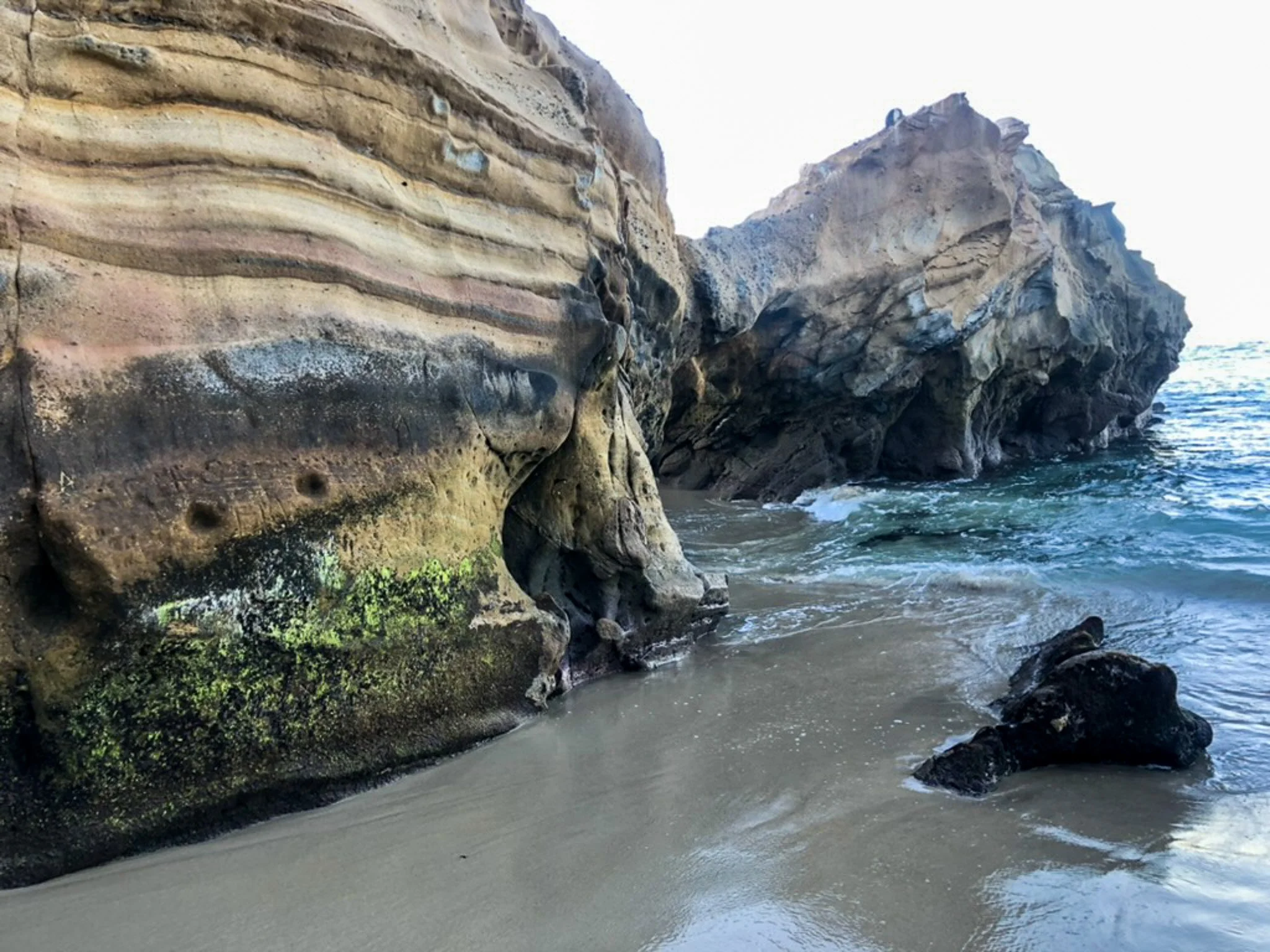 Rocky shoreline with layered cliffs, moss-covered rocks, gentle waves, and sandy beach