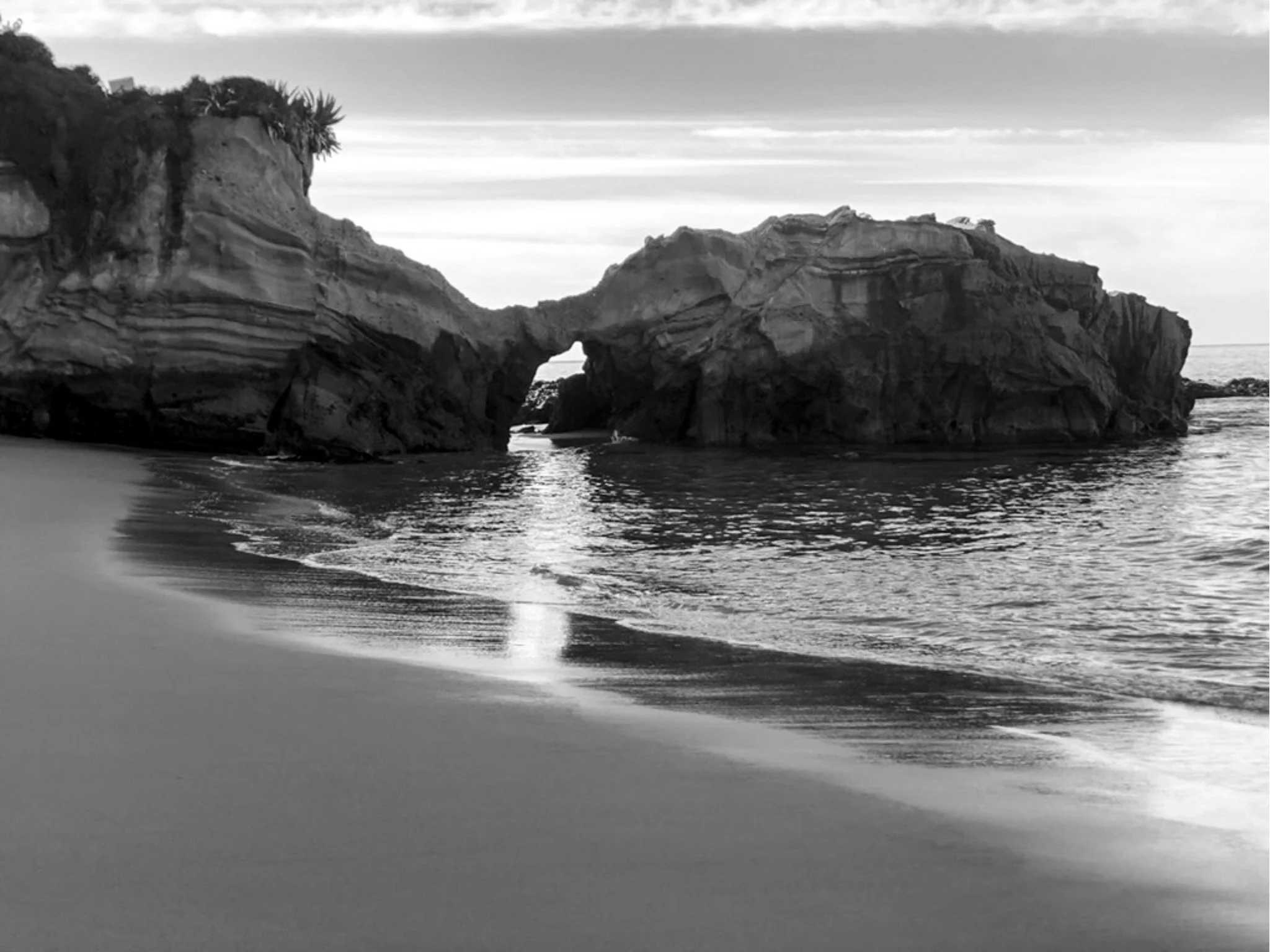 Black and white photograph of a beach with large rock formations, one of which has a natural arch, and the ocean in the background.