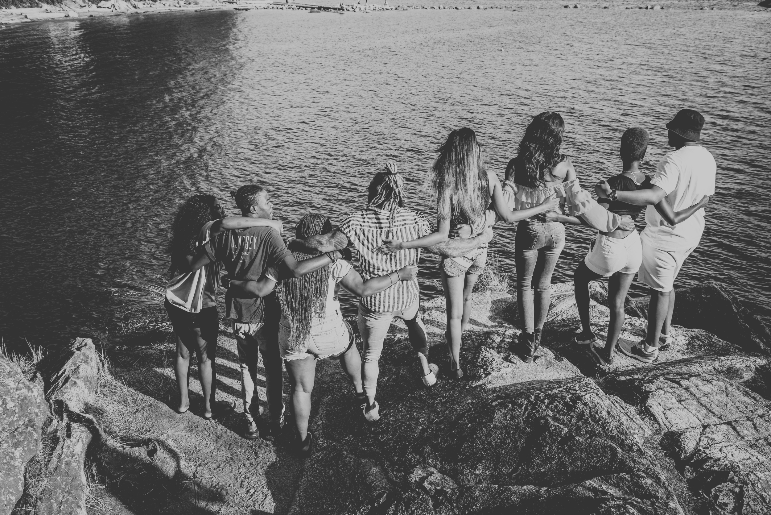 PNW Production. A group of friends standing on rocks, arms around each other, enjoying a scenic lake view.