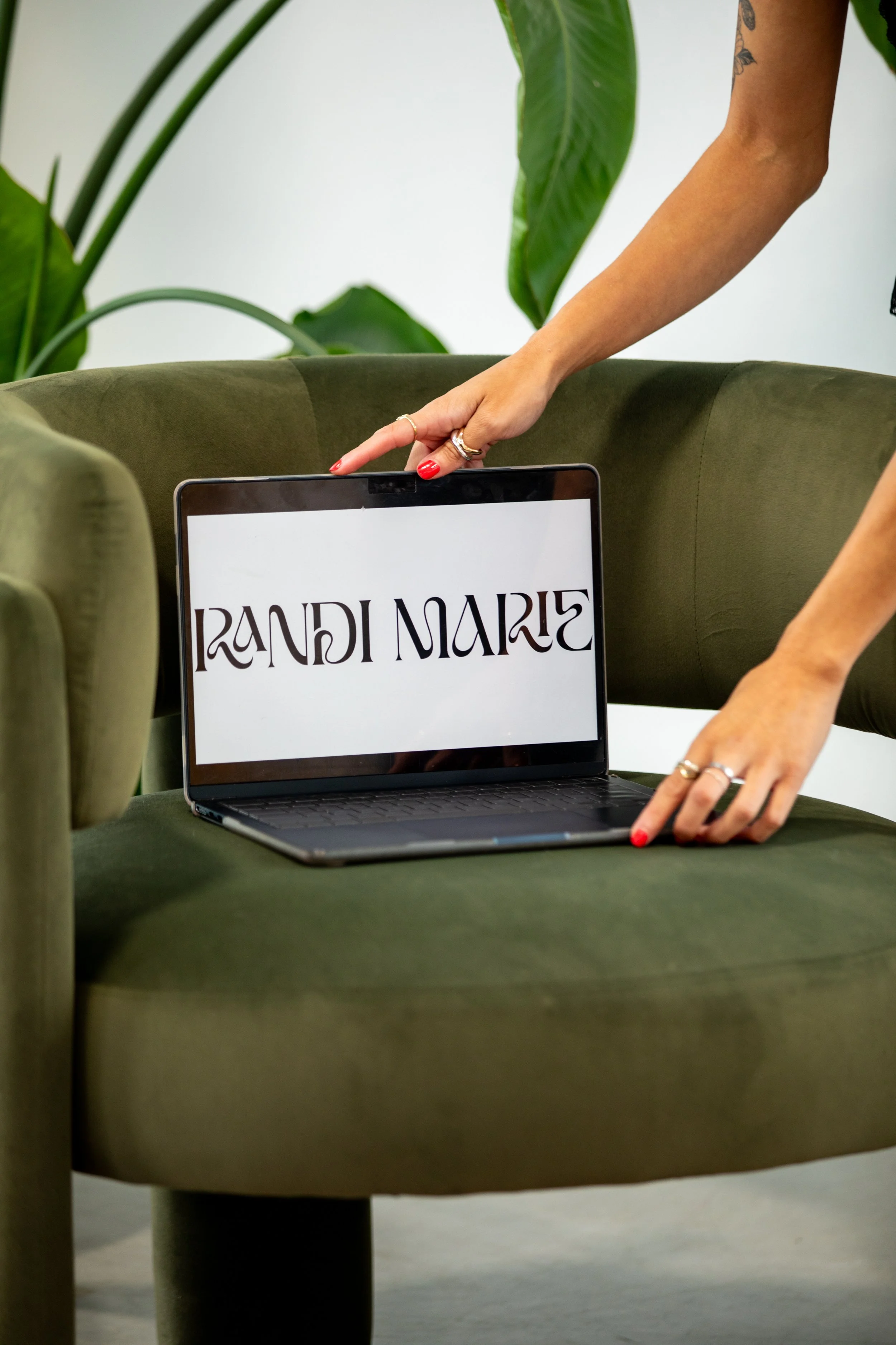 A person with painted nails and a ring on their finger is pointing at a laptop screen displaying the text 'Randi Marie.' The person's arm has a tattoo, and they are leaning over a green upholstered chair with large green leaves in the background.
