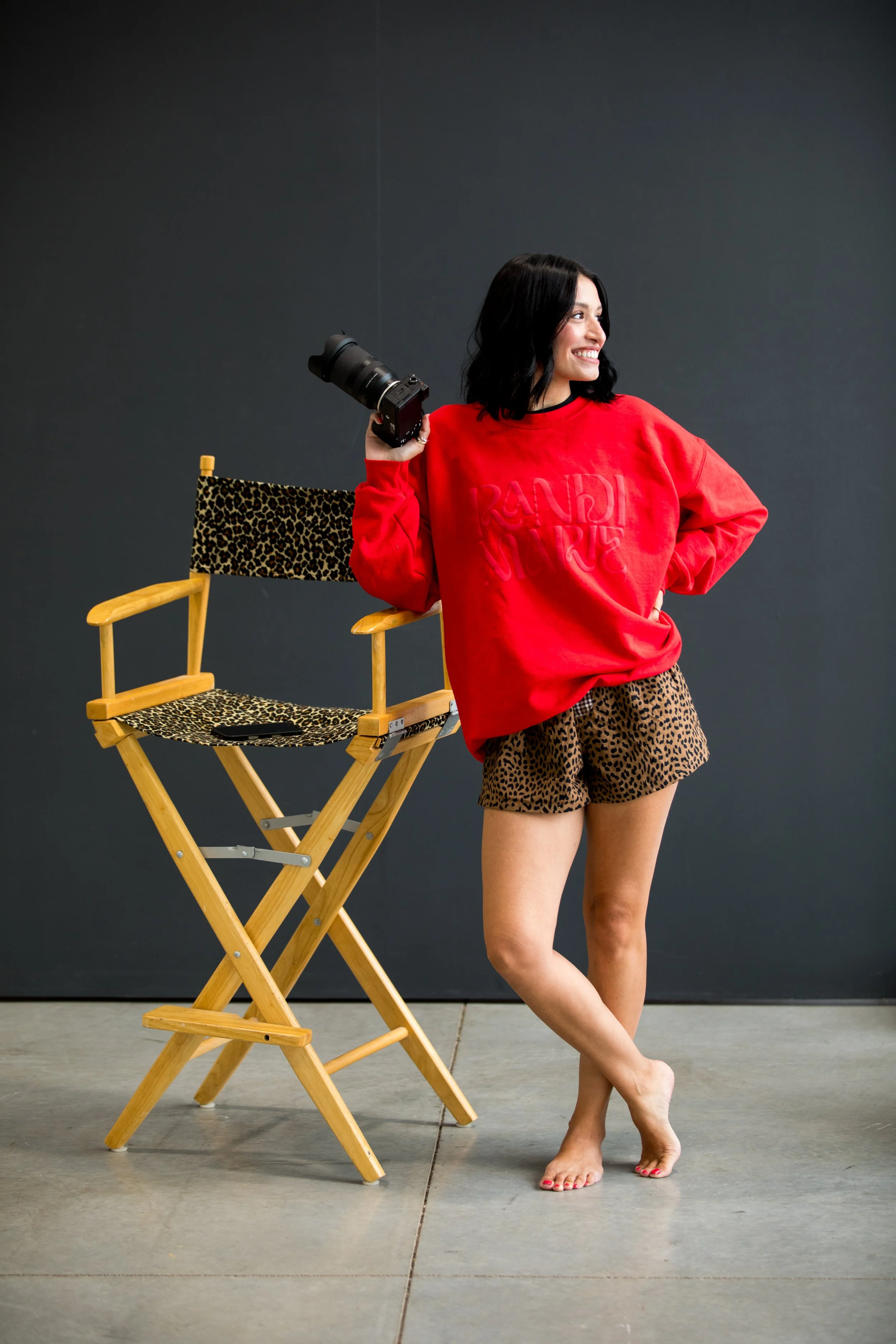 A woman with black hair smiling, standing barefoot with one hand on her hip, wearing a red sweatshirt and leopard-print shorts, next to a director's chair with leopard print fabric, holding a camera in her other hand, against a plain dark background.