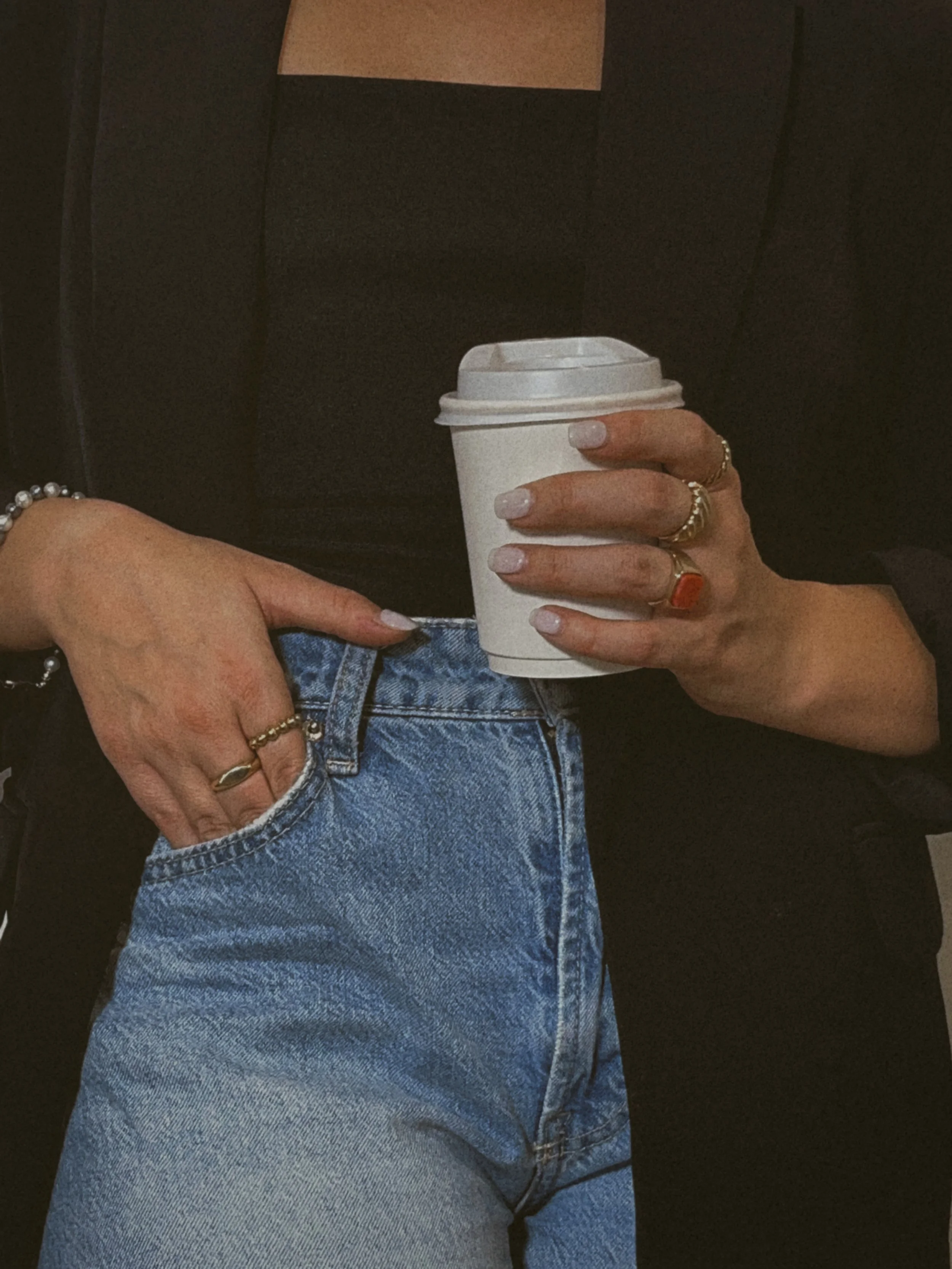 Person holding a white paper coffee cup with a lid, wearing blue jeans and a black top, with jewelry including rings and bracelets, and resting their hand on their hip.