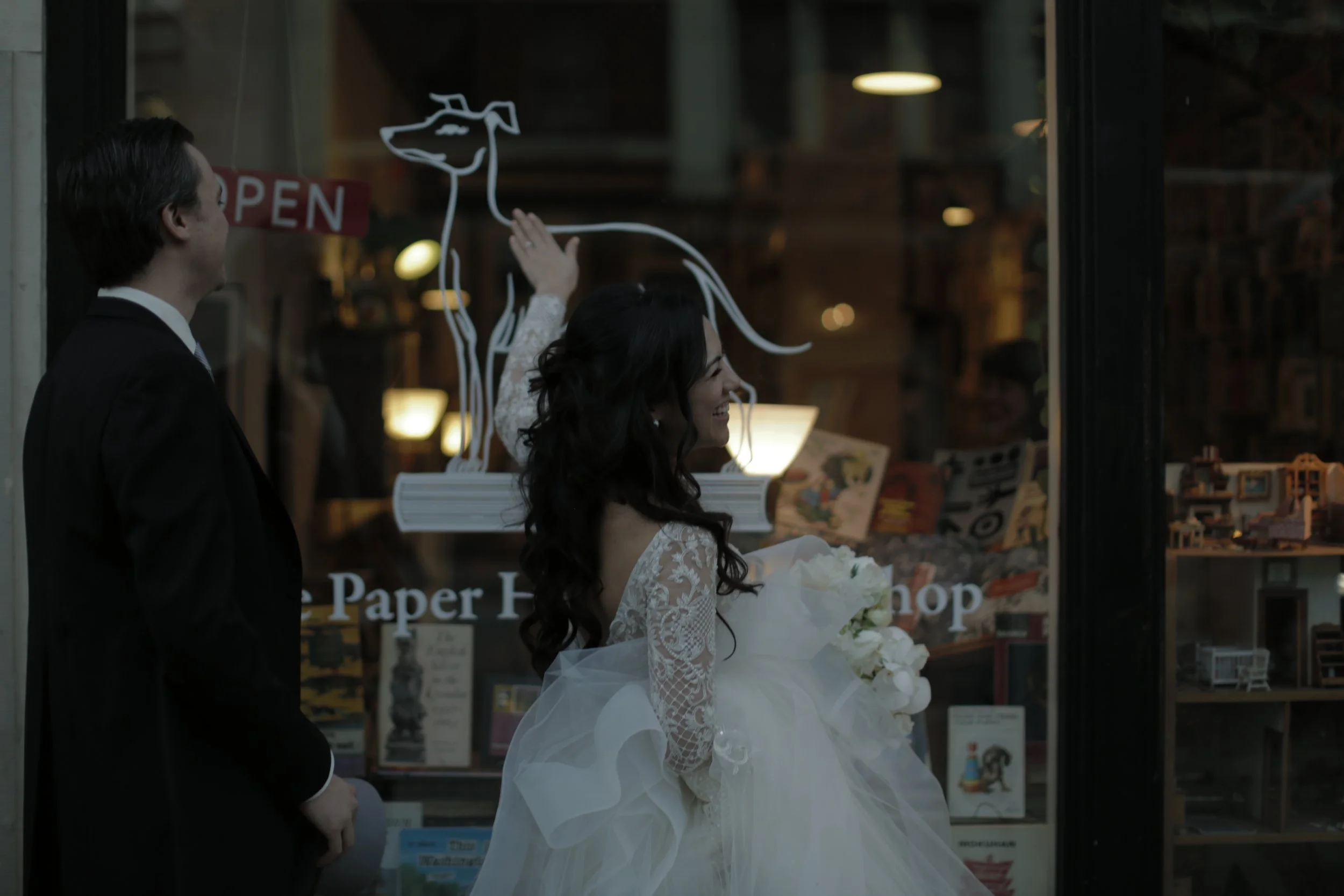 A bride in a white wedding dress and a groom in a black suit stand outside a shop window, smiling, with the bride holding a bouquet of white flowers.