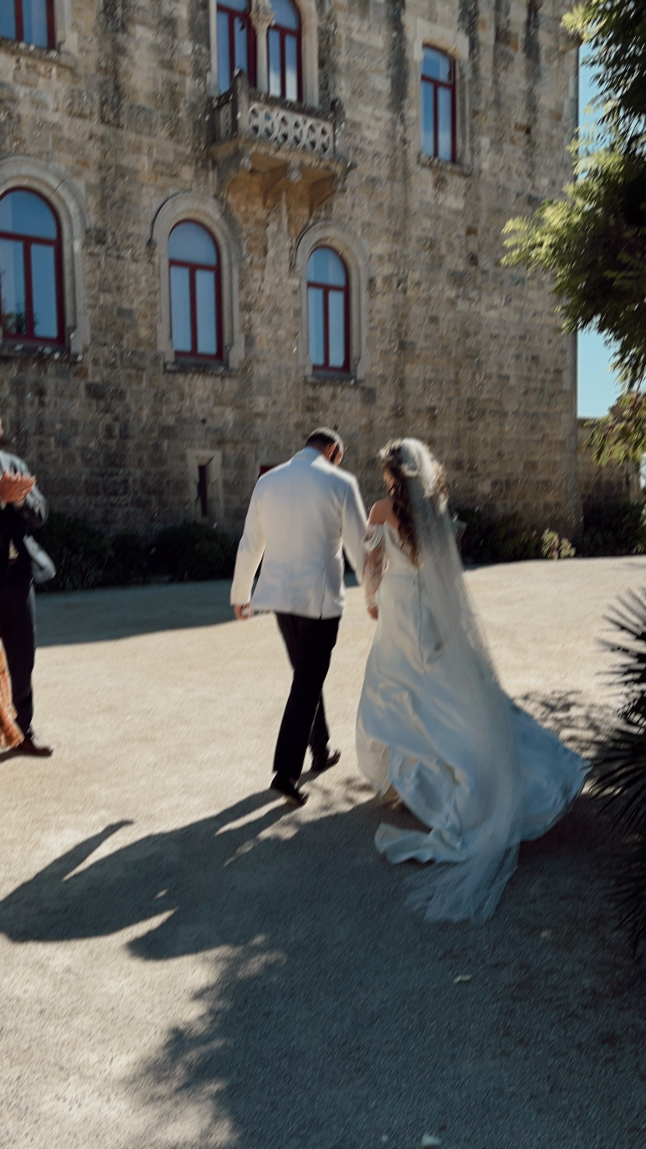 A bride and groom walking together outdoors near a stone building, with the bride wearing a white wedding gown and veil, and the groom in a white jacket and black pants. There are a few other people nearby and shadows cast on the ground.