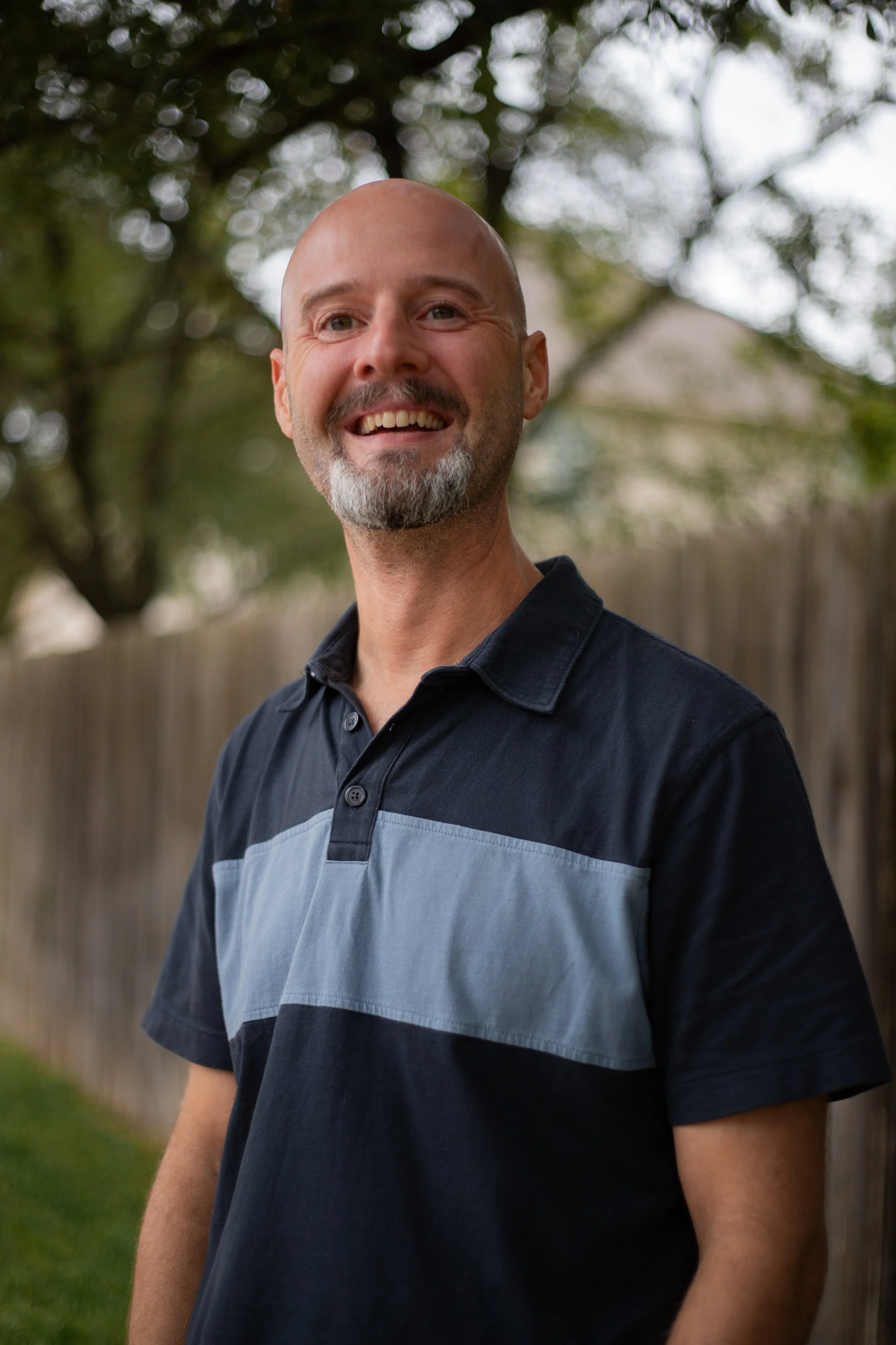 A smiling bald man with a beard, wearing a black and gray polo shirt, standing outdoors near a wooden fence and trees.