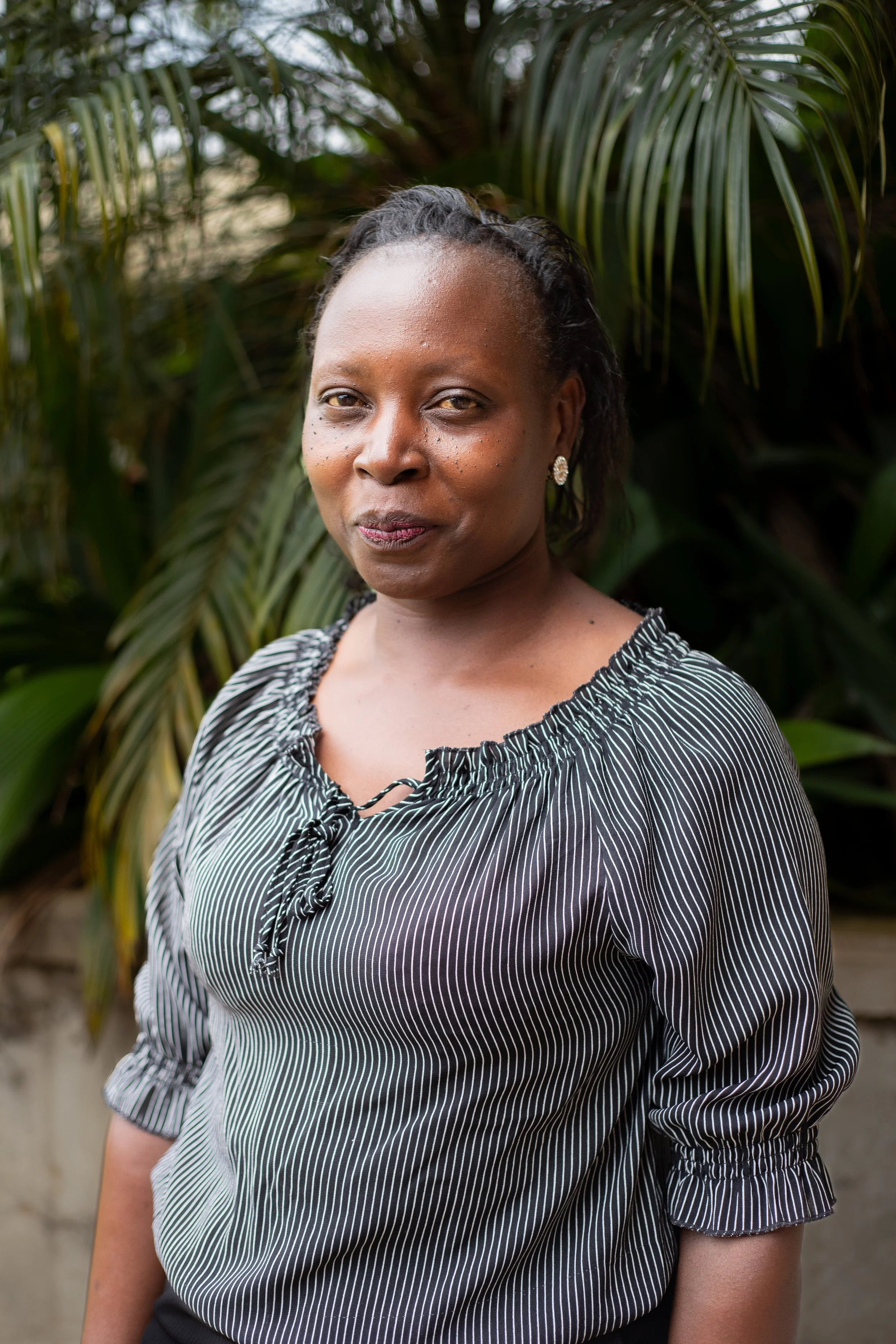A woman with short curly hair wearing earrings and a black-and-white striped blouse standing outdoors in front of green tropical plants.