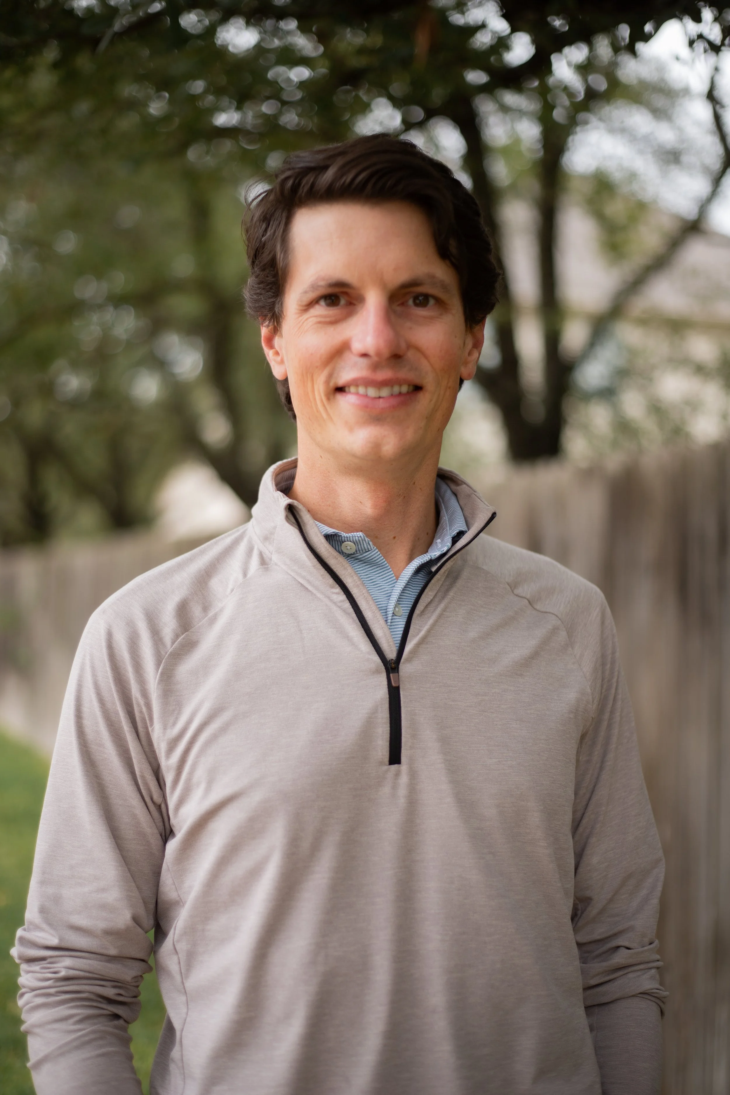 A man standing outdoors in front of trees and a wooden fence, wearing a beige quarter-zip pullover and smiling.