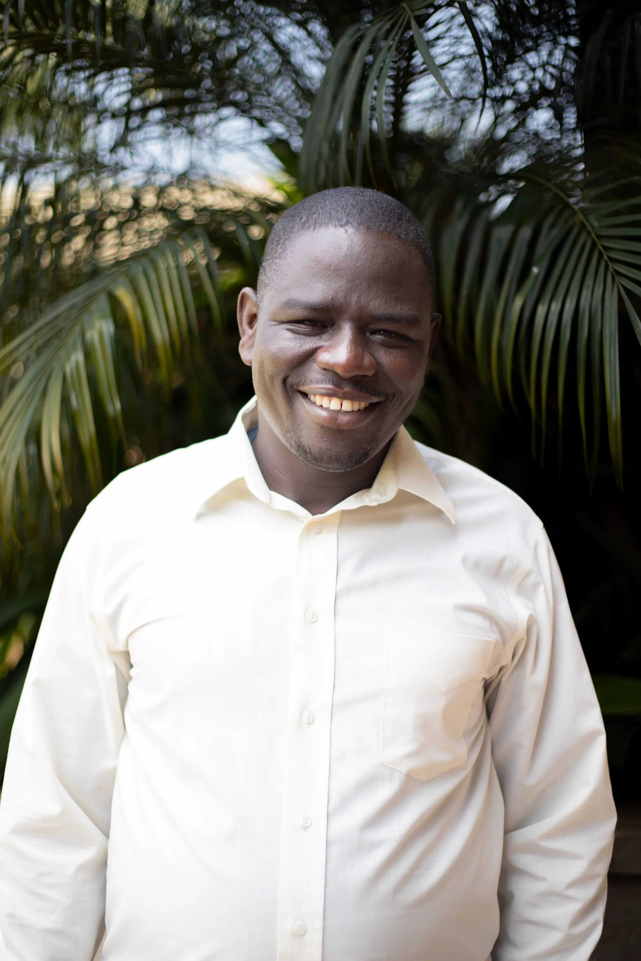 A smiling man in a white dress shirt standing outdoors in front of green foliage.
