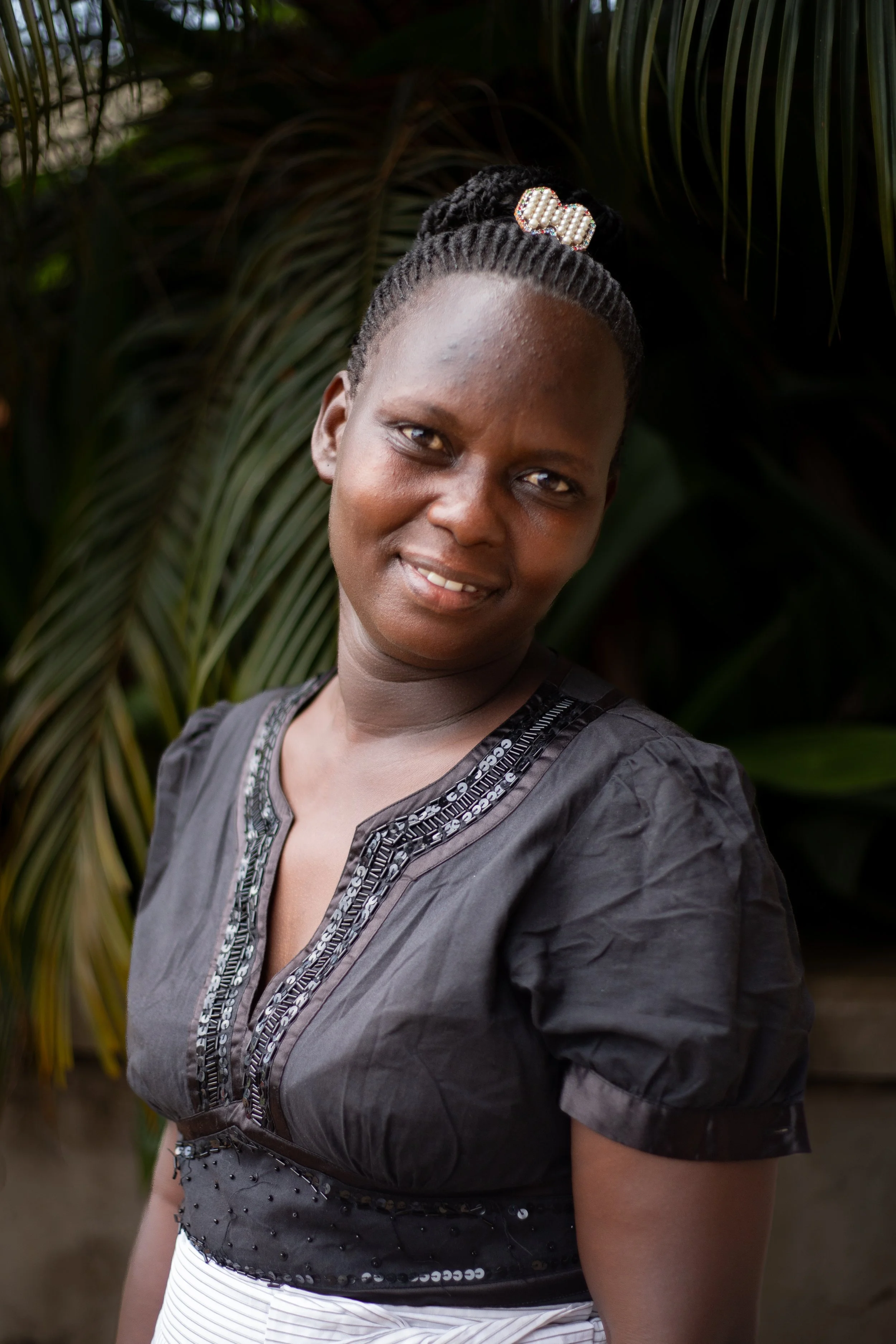 Portrait of a smiling woman with braided hair and a hair accessory, wearing a black top with decorative trim, standing outdoors against a background of green foliage.