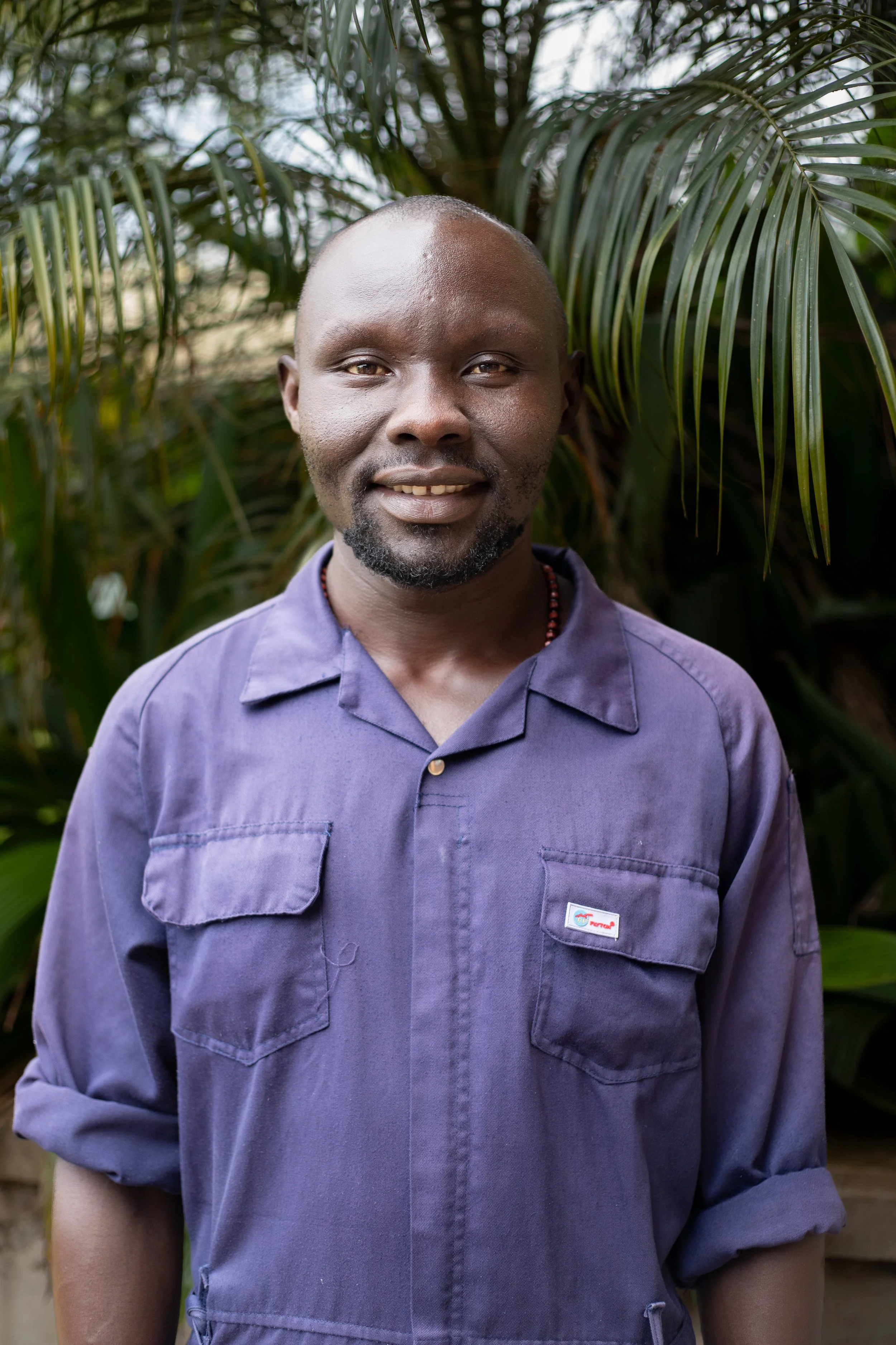 Portrait of a smiling man with a trimmed beard, wearing a purple work jumpsuit, standing outdoors in front of green tropical plants.