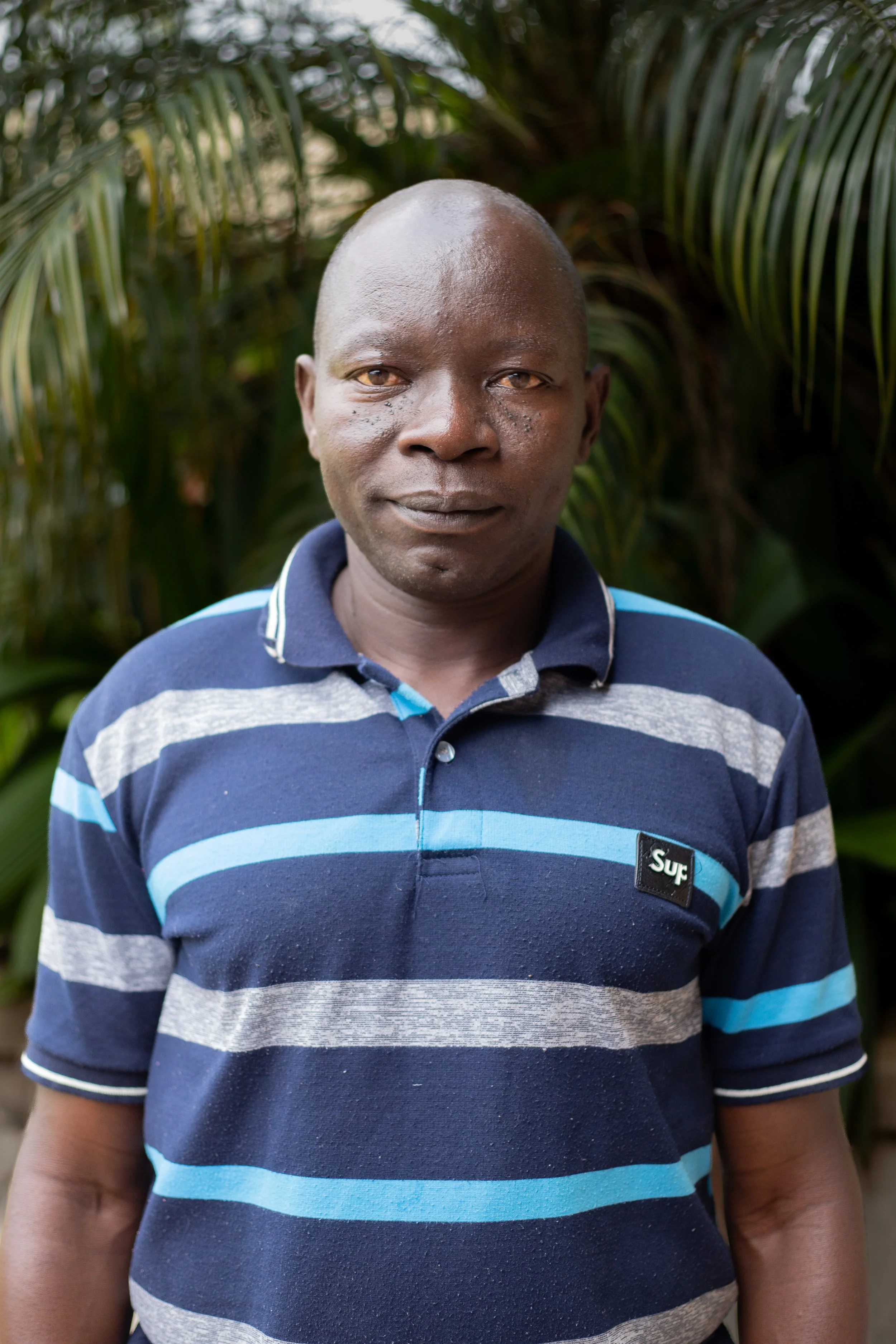 A middle-aged man with dark skin and a shaved head standing outdoors in front of green foliage, wearing a navy blue, gray, and light blue striped polo shirt.