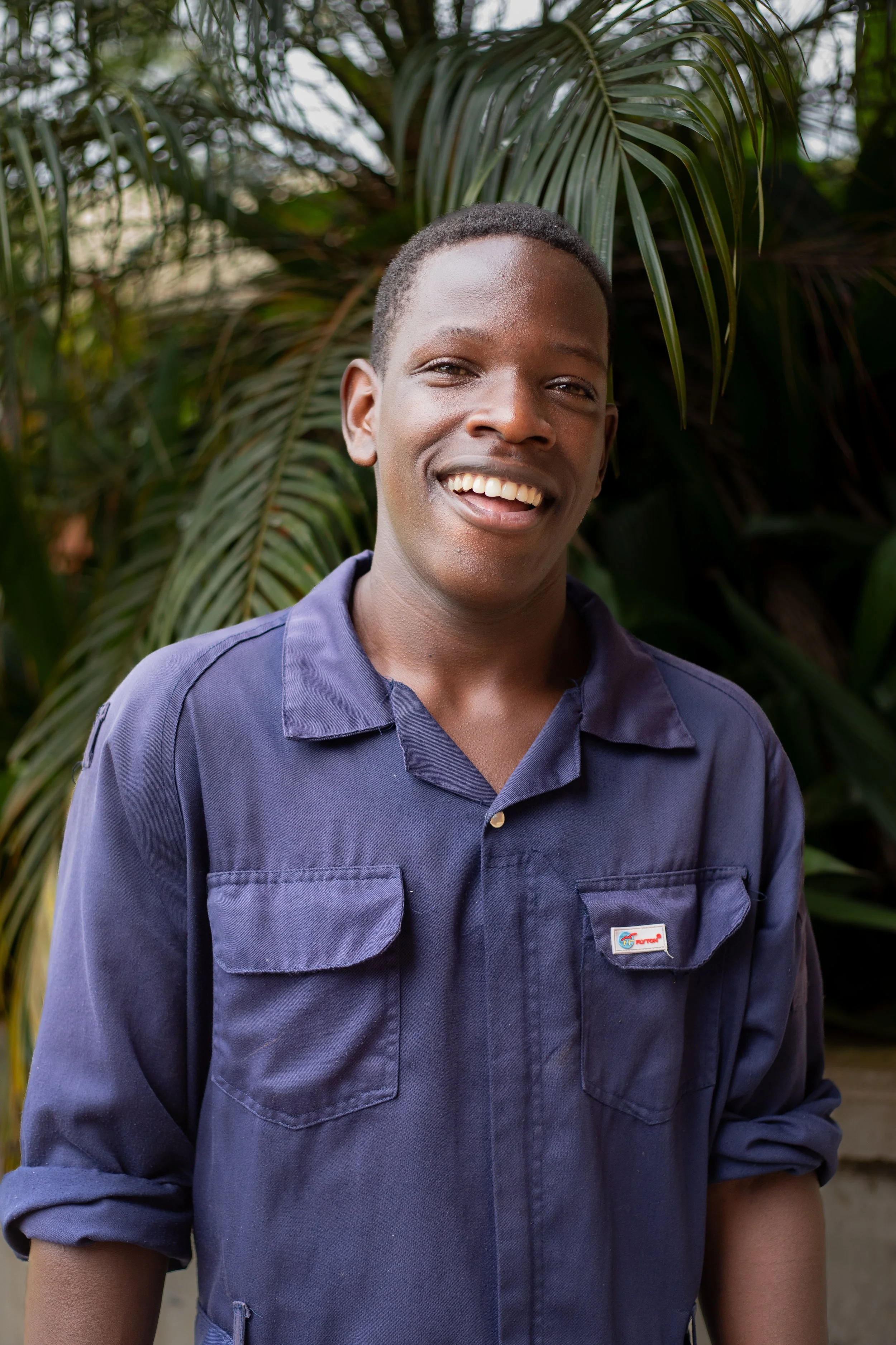 A young man with short hair wearing a navy blue shirt, smiling, standing outdoors in front of lush green foliage.