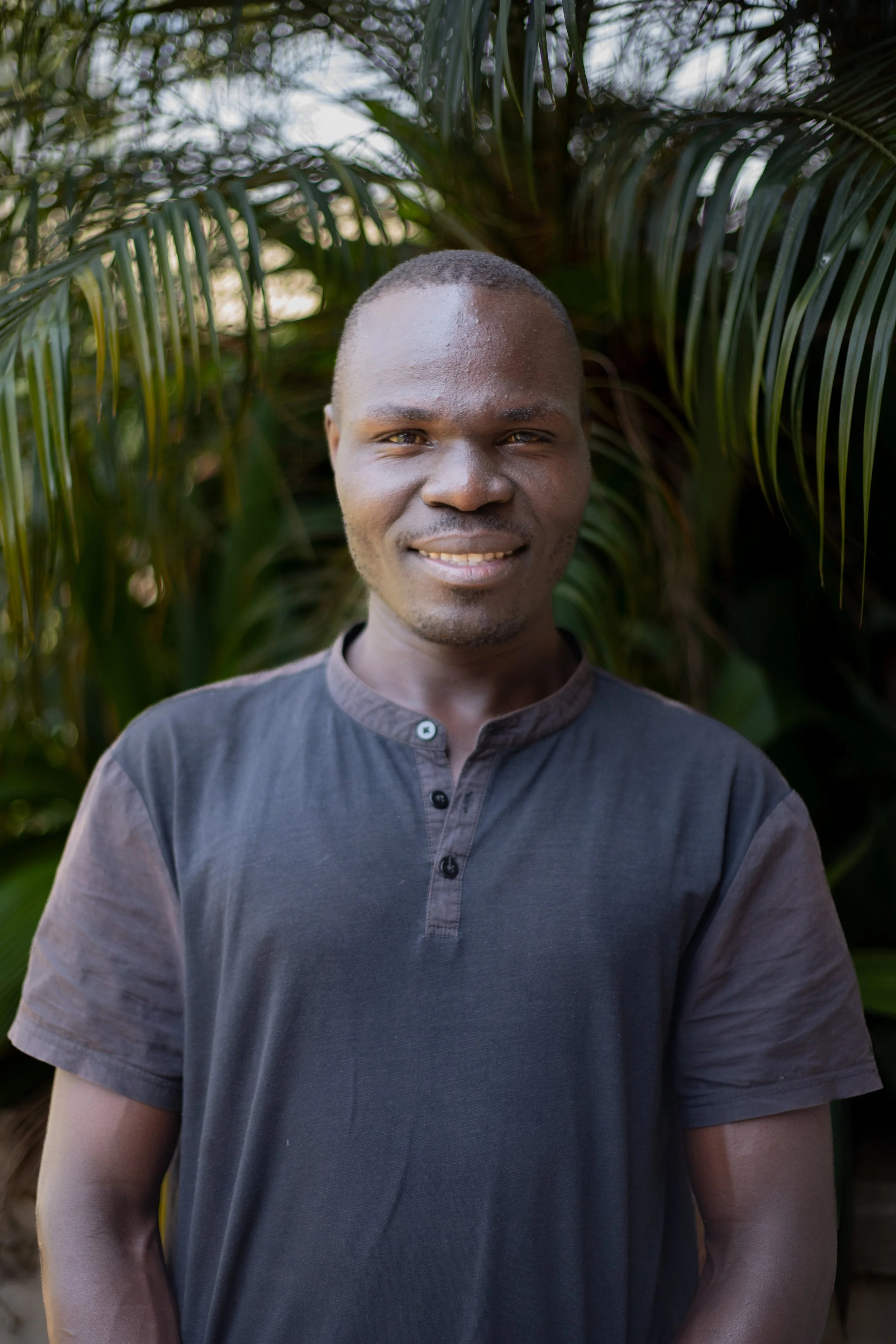A man standing outdoors in front of green palm leaves, smiling at the camera.
