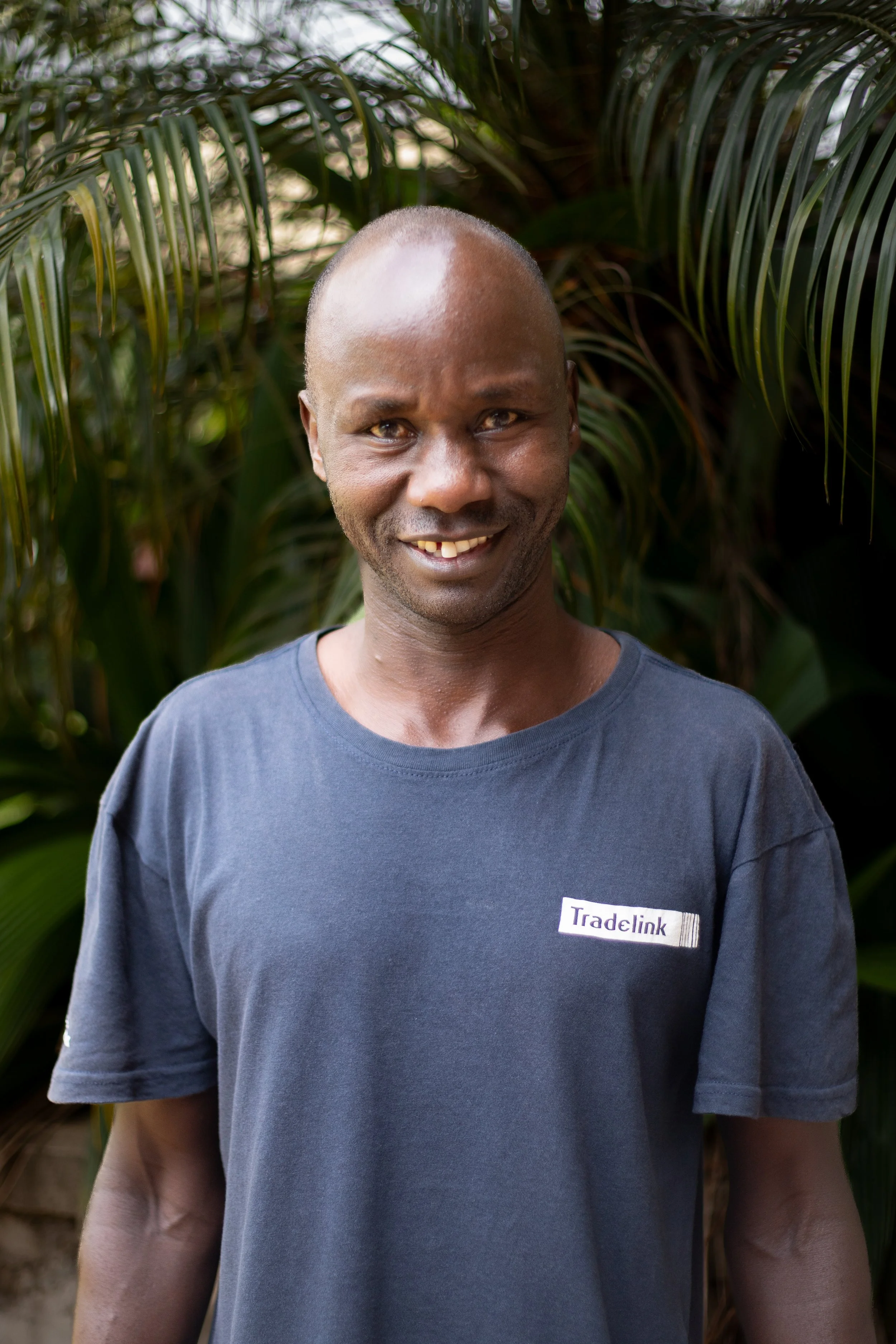 A smiling man with a shaved head and dark skin stands outdoors in front of lush green plants, wearing a dark T-shirt with a 'Tradelink' logo.