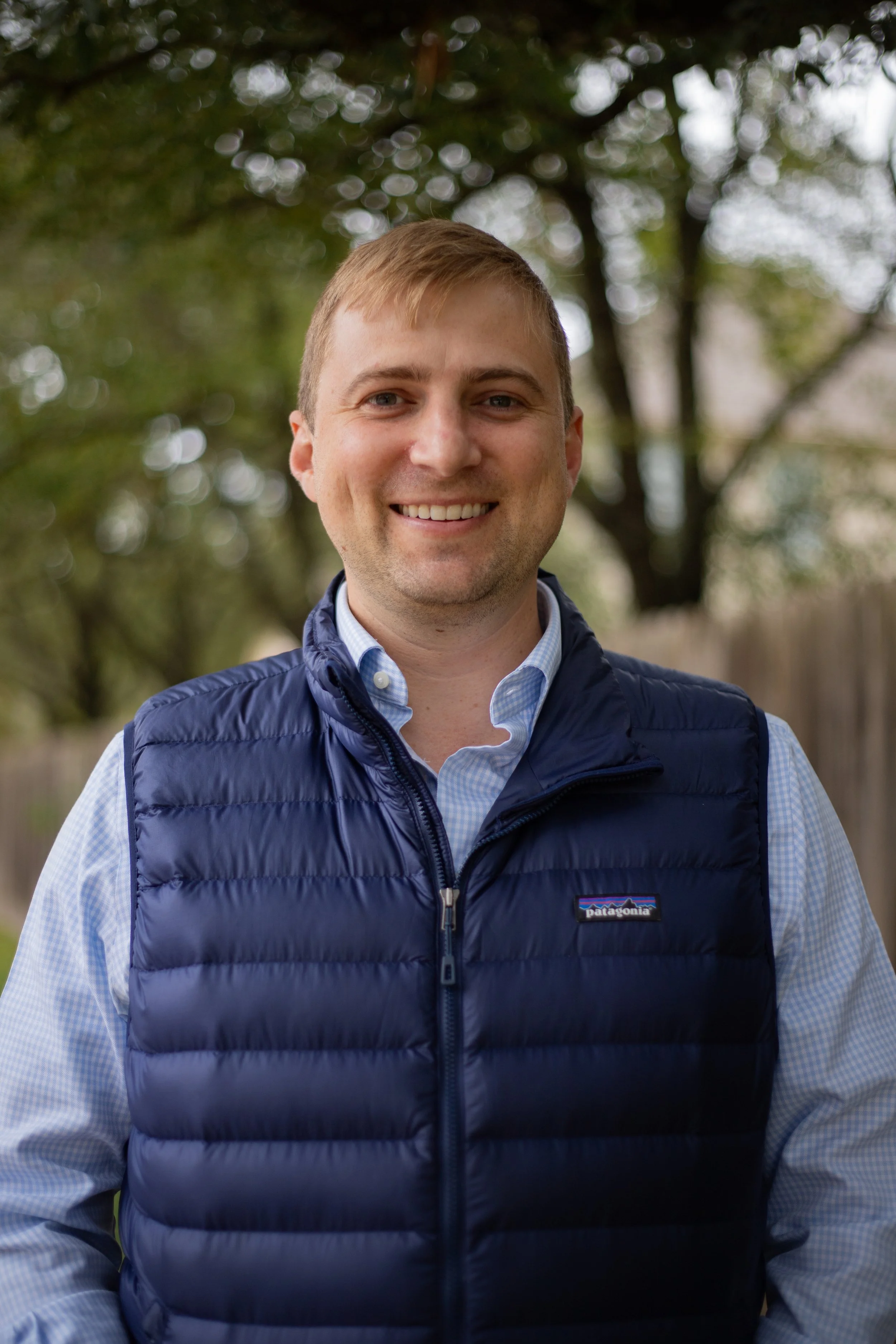A young man with light brown hair smiling outdoors, wearing a blue quilted Patagonia vest over a checkered button-up shirt, standing in a wooded area.