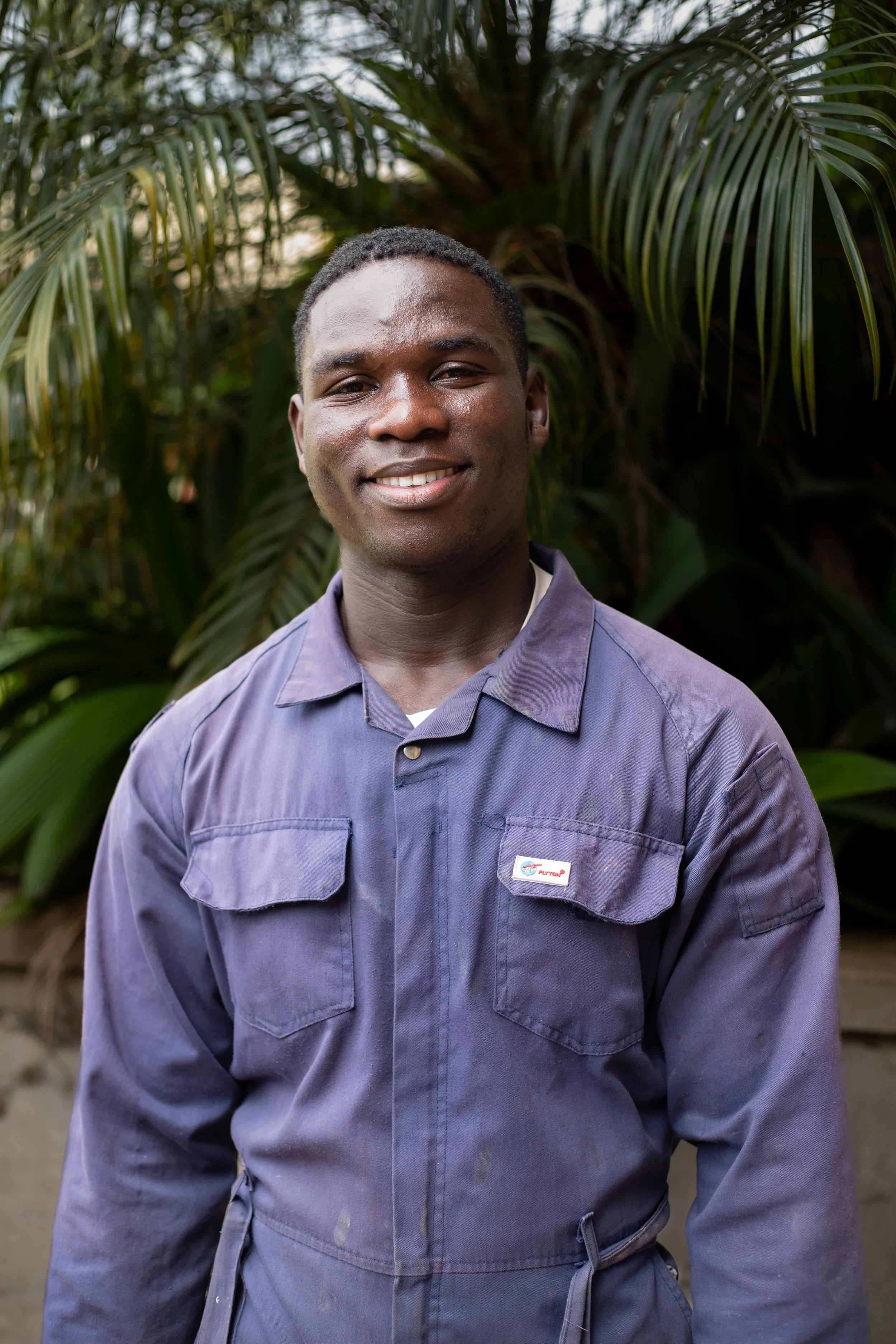 A young man in a work jumpsuit standing outdoors in front of green plants, smiling at the camera.