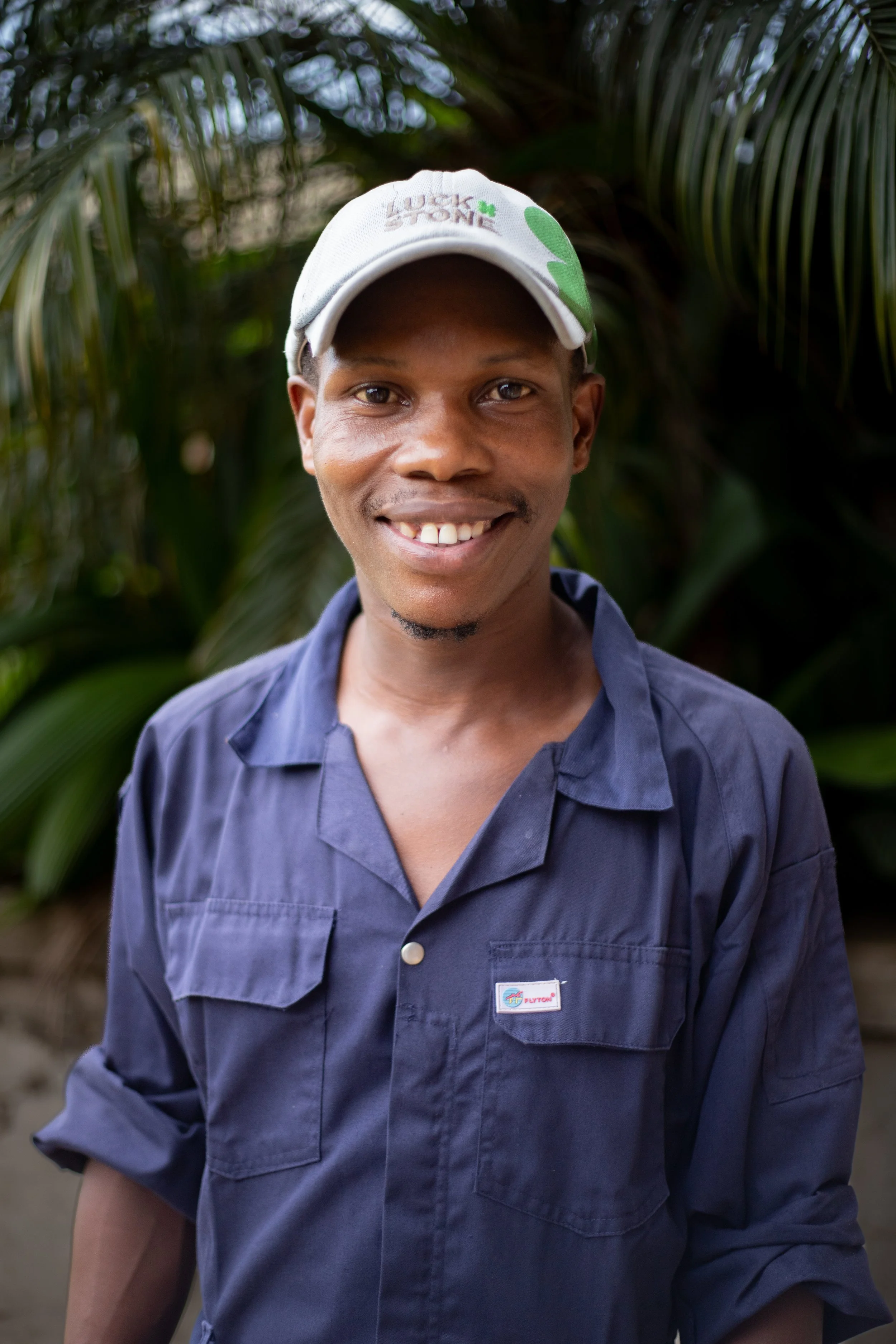 Smiling man wearing a baseball cap and navy blue work shirt outdoors with green foliage in the background.