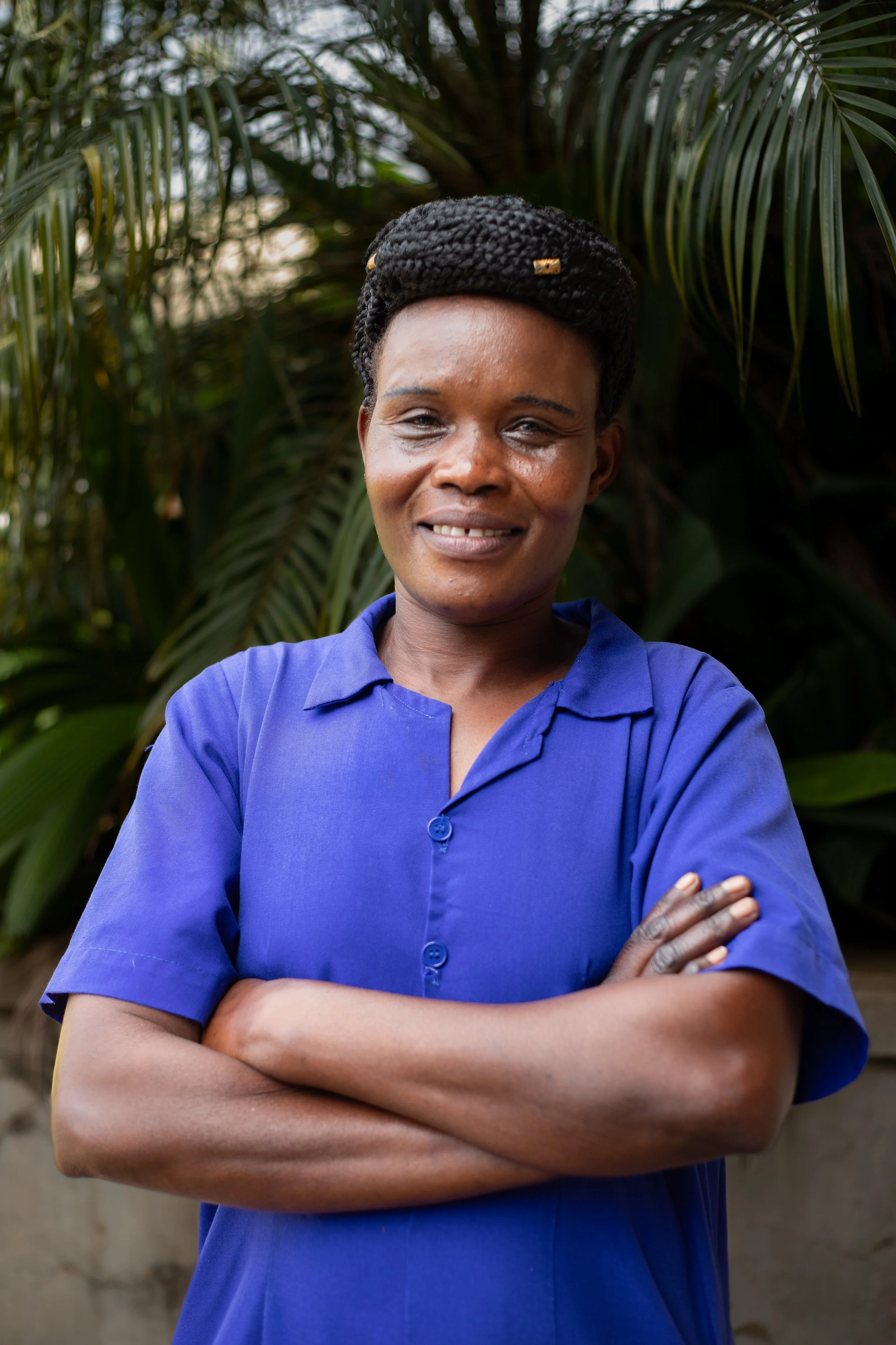 A woman with dark skin and braided hair standing outdoors with crossed arms, smiling, wearing a blue shirt, in front of green tropical plants.