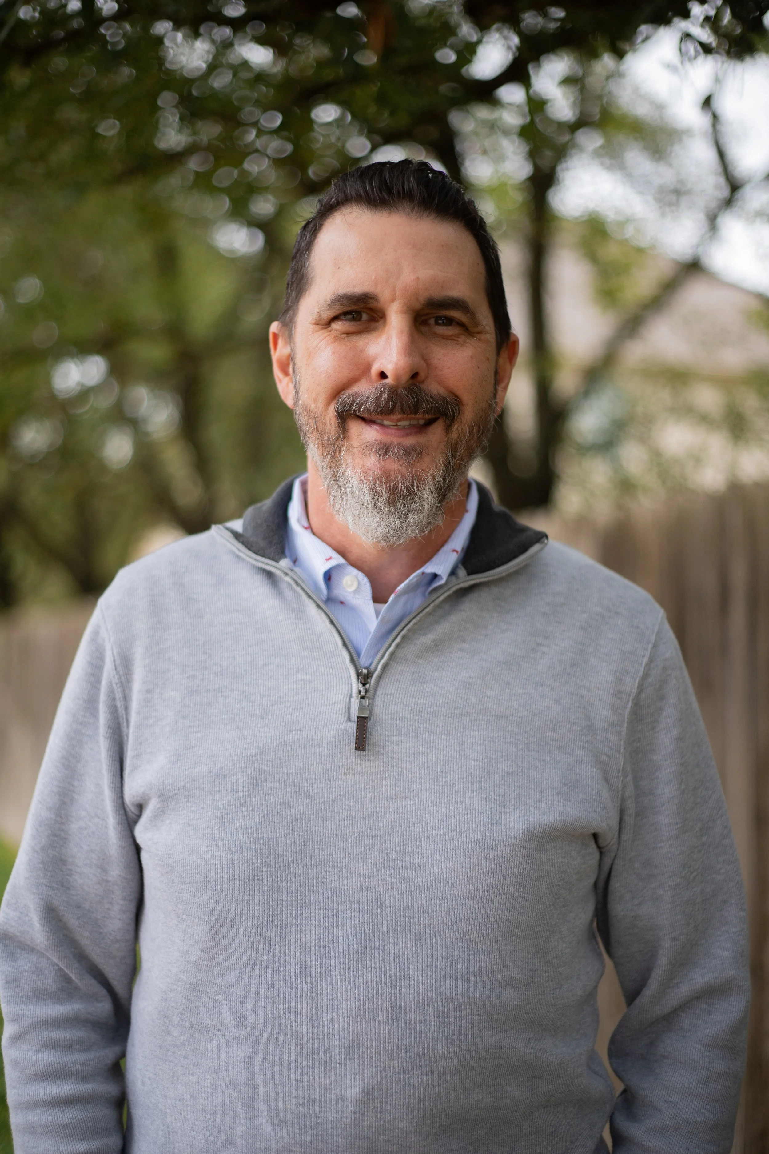 A smiling middle-aged man with dark hair, a beard, and a mustache, wearing a gray zip-up sweater over a collared shirt, standing outdoors with trees in the background.