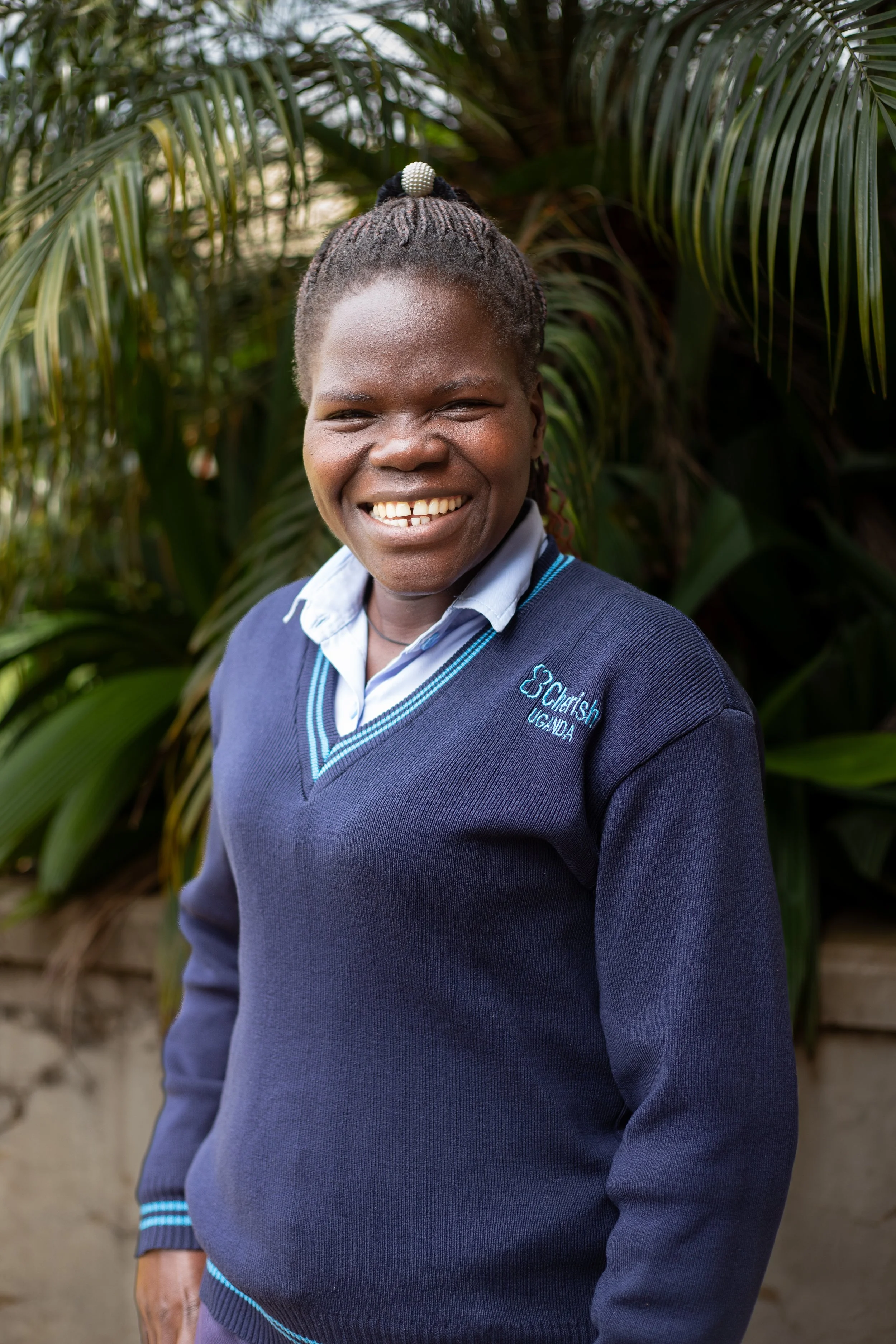 A smiling woman wearing a school uniform, standing outdoors with greenery in the background.