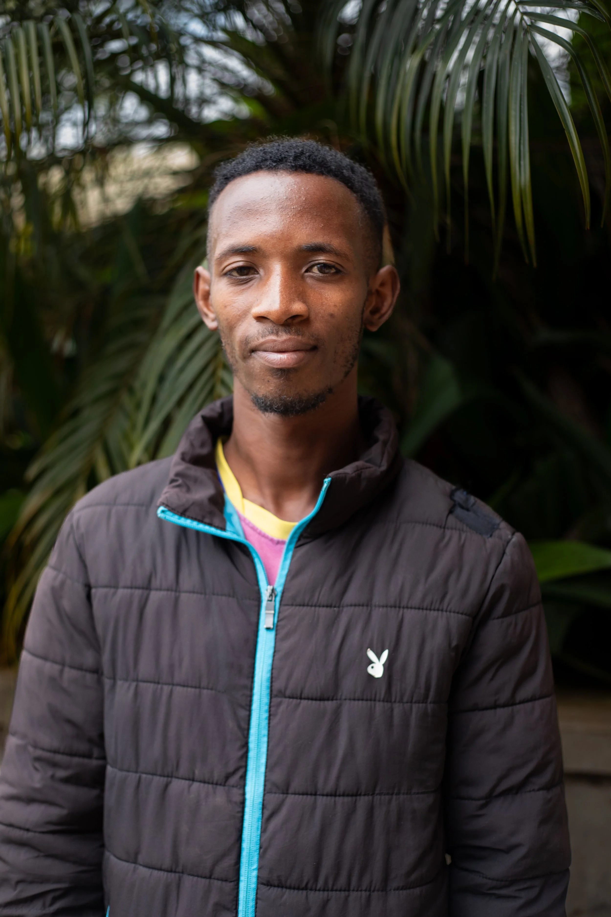 A young man with short curly hair and a slight beard standing outdoors in front of green tropical plants, wearing a black jacket with bright blue zipper details.