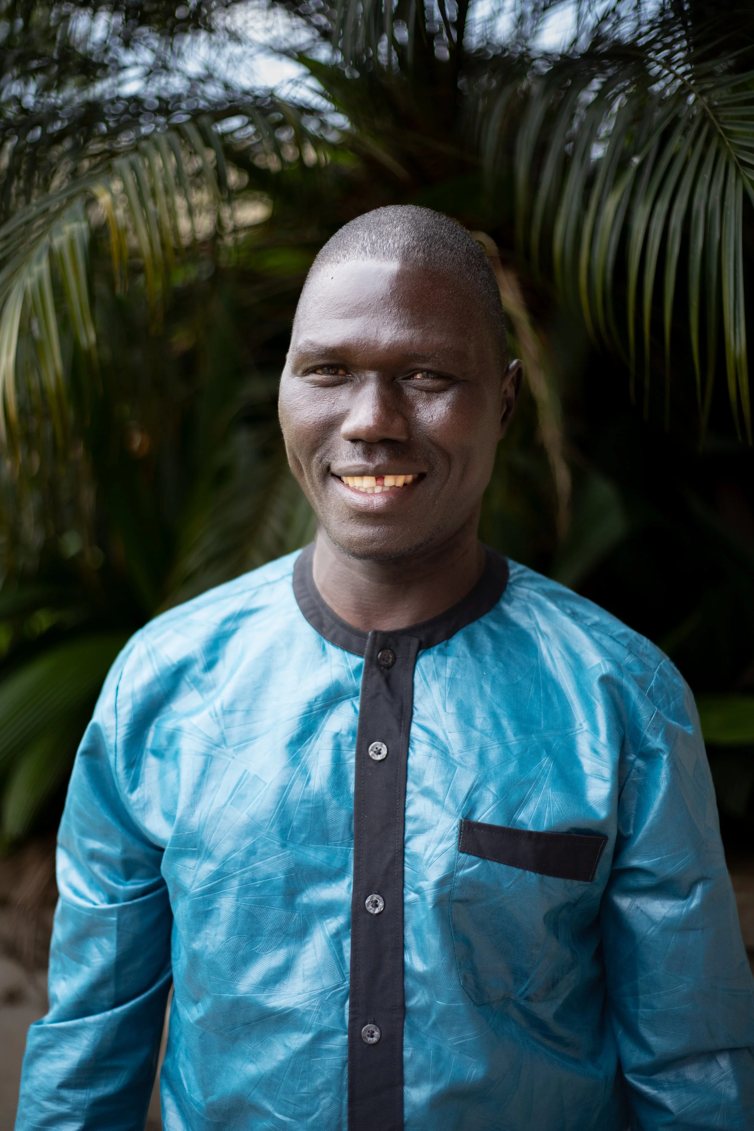 A smiling man with dark skin and short hair, wearing a shiny blue shirt with black trim and buttons, standing outdoors in front of lush green foliage.