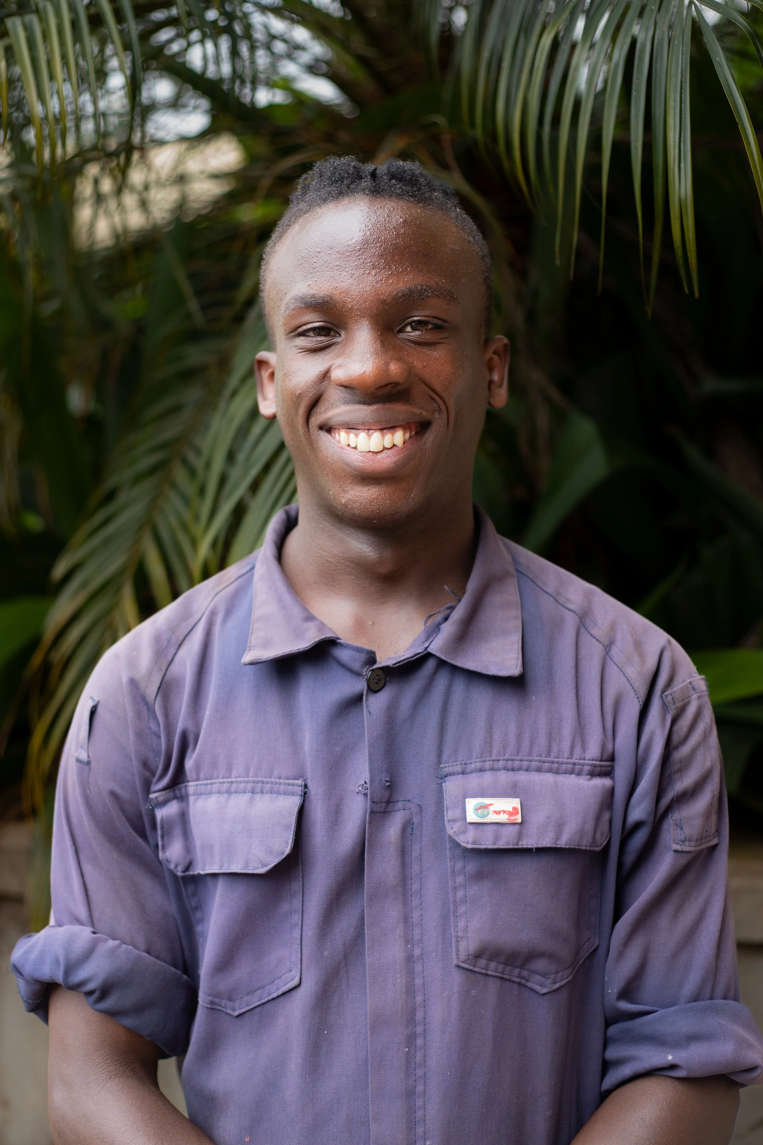 A young man with short dreadlocks smiling outdoors with tropical plants in the background, wearing a purple button-up shirt with rolled sleeves.