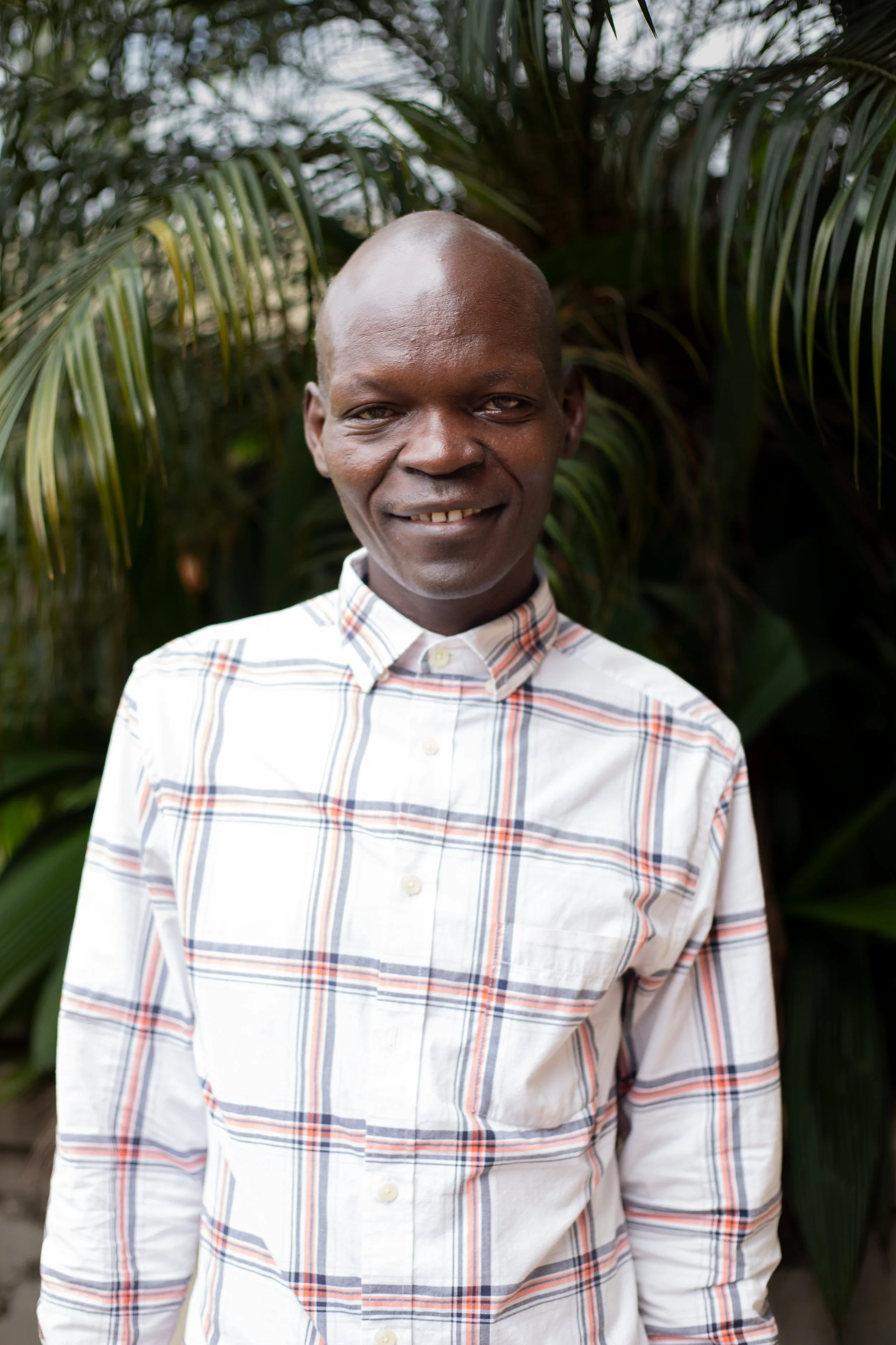 A smiling man with dark skin, wearing a white plaid shirt with red, blue, and orange stripes, standing outdoors in front of green foliage.