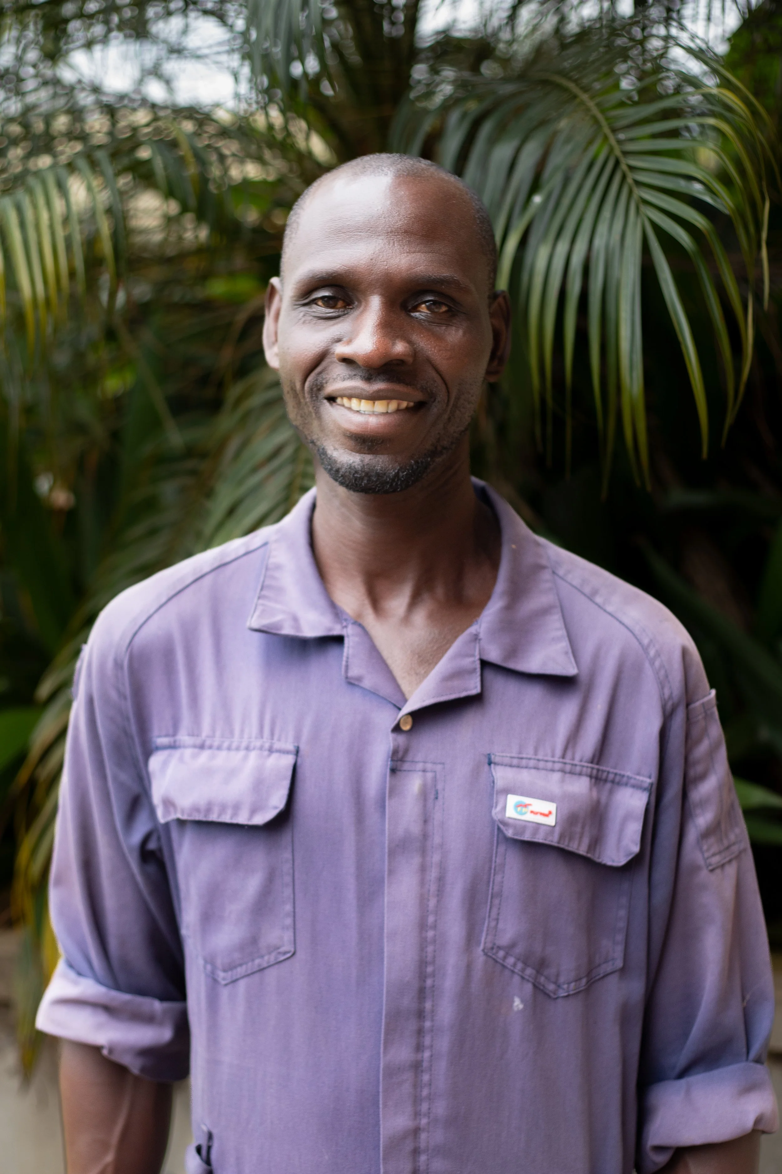 A smiling man in a purple shirt standing outdoors with green tropical plants in the background.