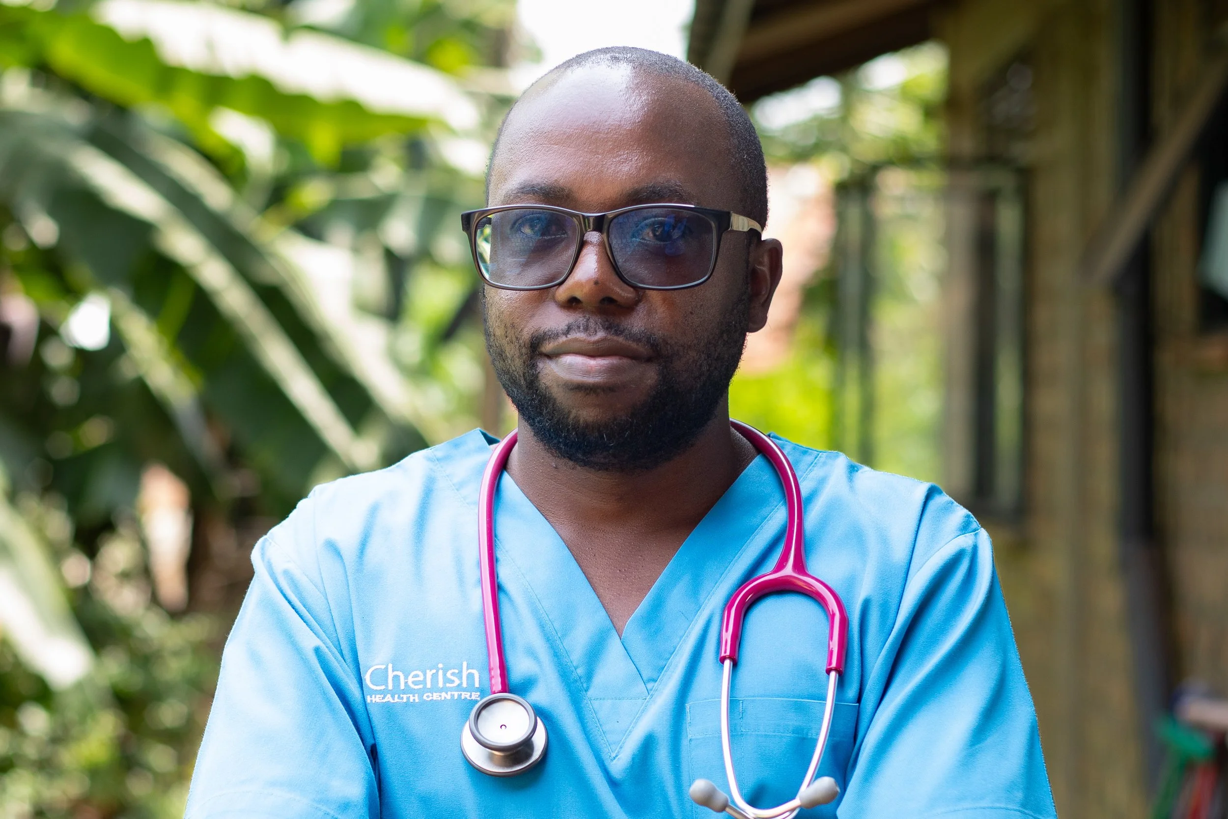 A smiling man wearing a white medical coat with 'Cherish Health Centre' logo, and light green scrubs, standing outdoors with lush green plants in the background.