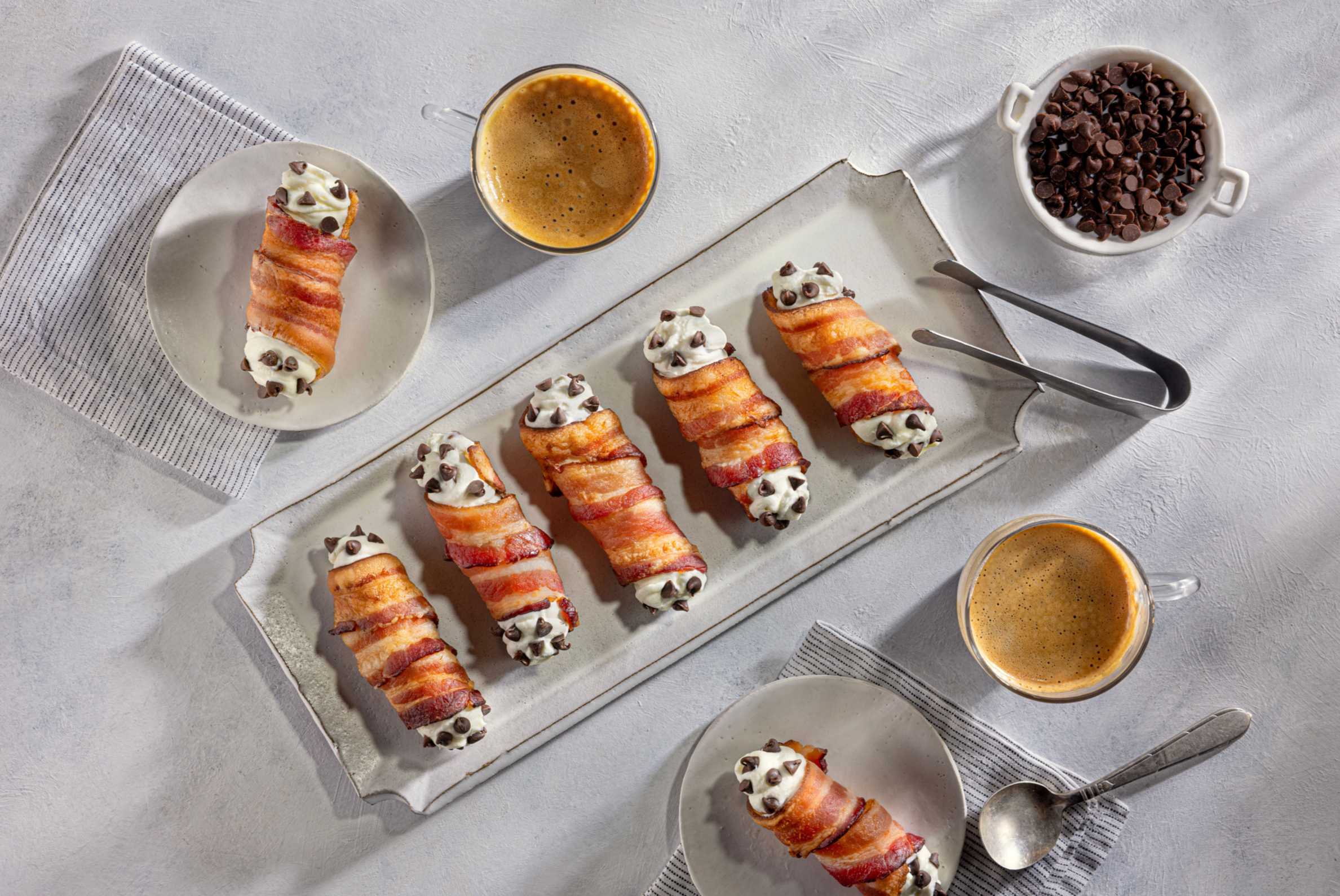 Bacon-wrapped snacks topped with cream and chocolate chips on a large rectangular platter, with cups of coffee and small plates of the same snacks on a light table.