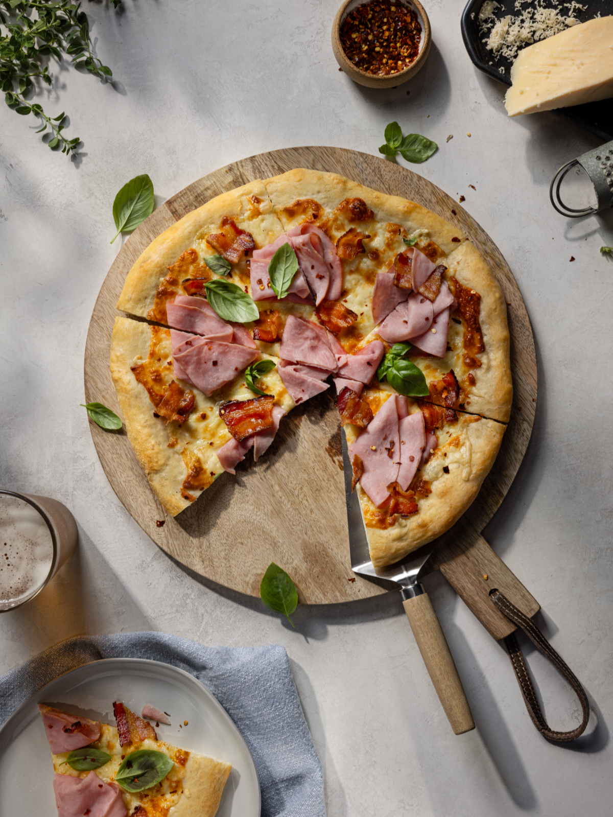 A pizza with ham and bacon on a round wooden pizza peel, with one slice being cut. There is a plate with a slice of pizza, a small bowl of red chili flakes, and a block of cheese nearby. Green basil leaves are garnished on the pizza.