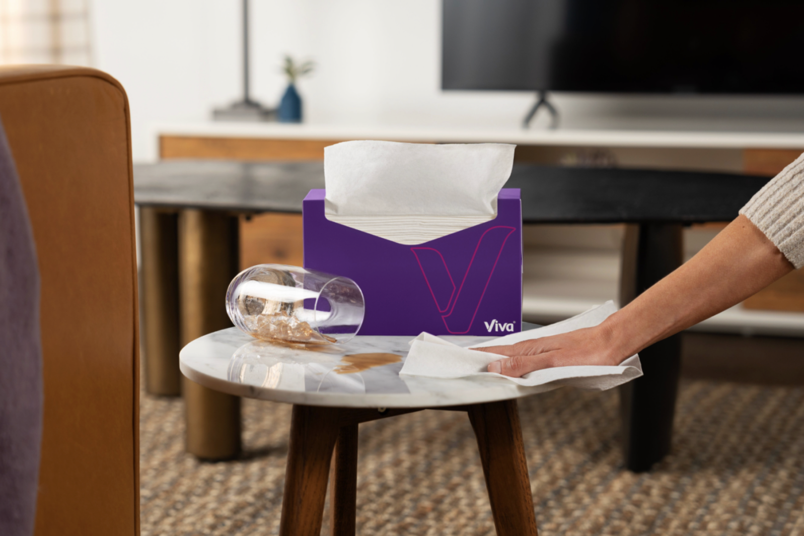 A purple tissue box labeled 'Viva' on a small round marble-top table, with tissue paper partially pulled out and a clear glass with some coins and a cloth inside, in a living room setting.