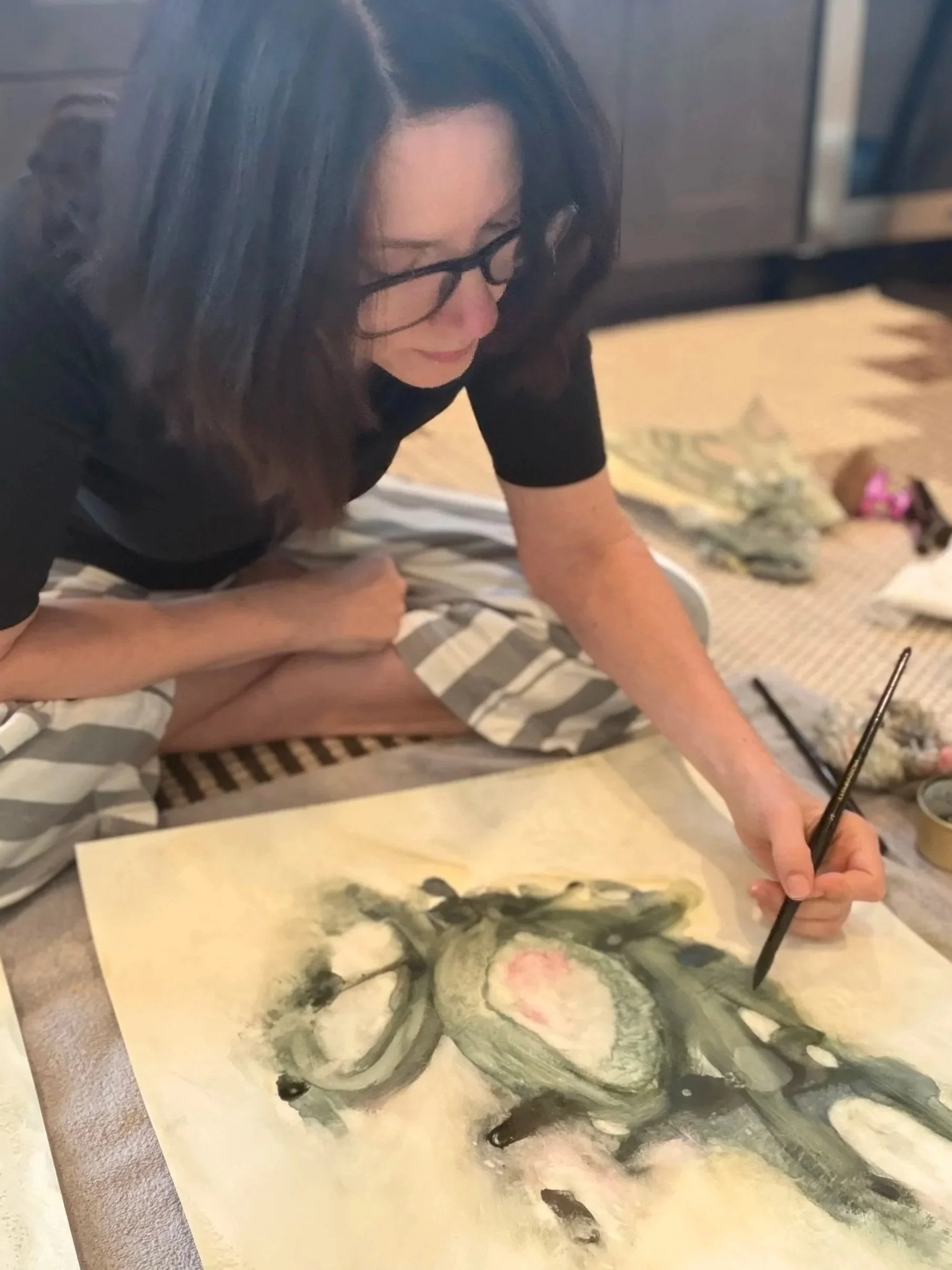 Angela Justice kneeling on the floor, painting an abstract watercolor with a fine brush, close-up view of hand and paper