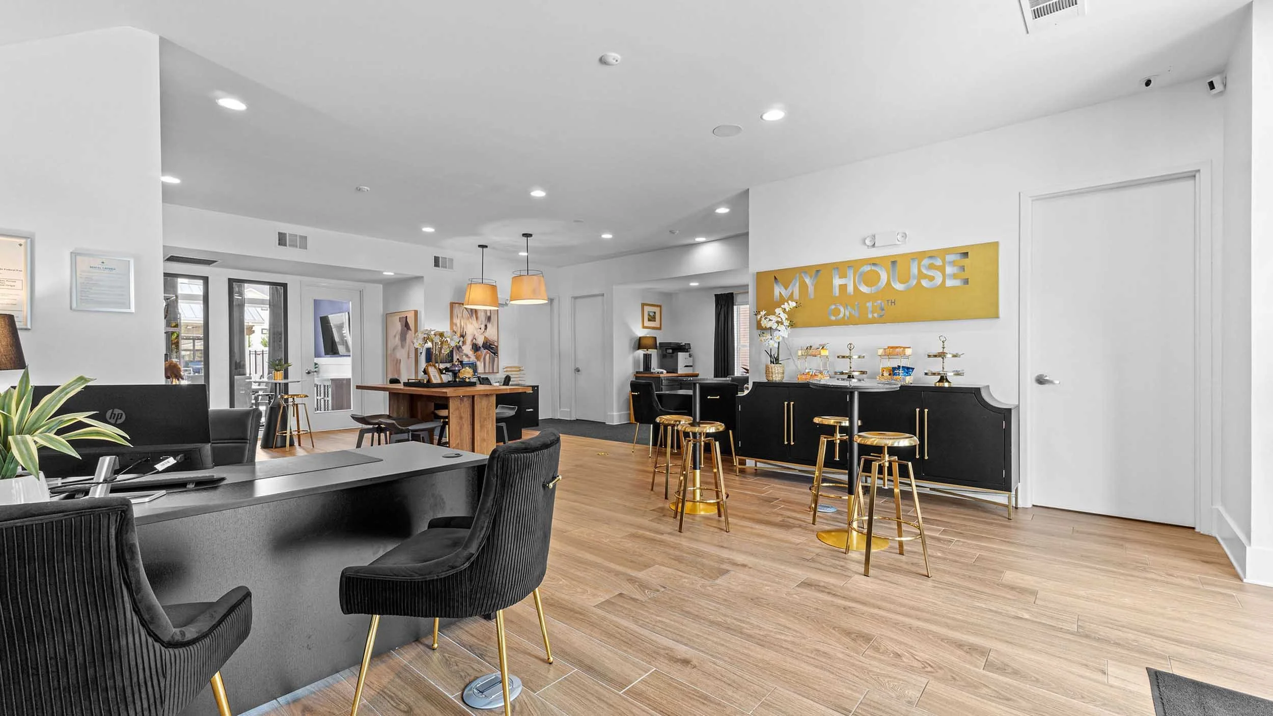Interior of a modern lounge with a reception desk, seating area with black and gray chairs, wooden flooring, and a large yellow sign reading 'MY HOUSE ON 13TH'. There are pendant lights, artwork on the walls, and a refreshment bar with gold stools.