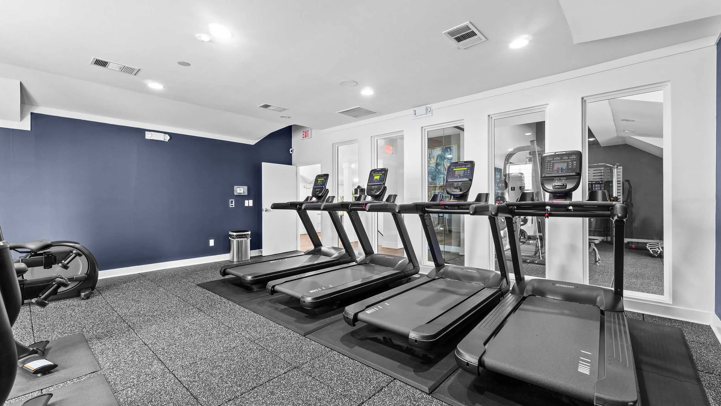 A fitness room with several treadmills and elliptical machines positioned in front of a glass-walled room with gym equipment inside, white walls, a dark blue accent wall, and a gray carpeted floor.