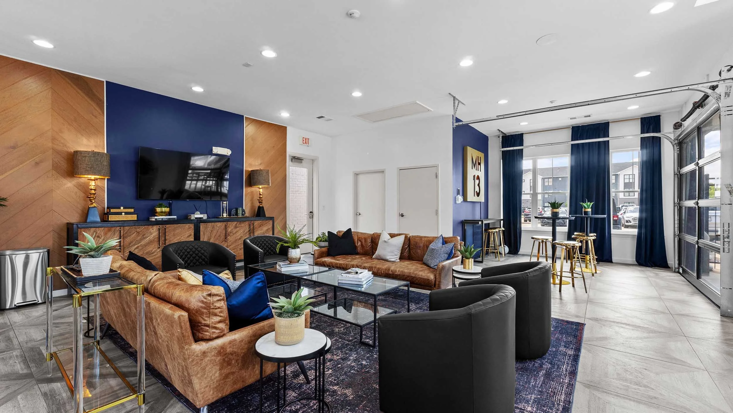 A modern interior lounge with a wood-paneled accent wall, a large flat-screen TV, and various comfortable seating options including leather and fabric sofas and chairs. The space is decorated with small potted plants, cushions, and tables, with large
