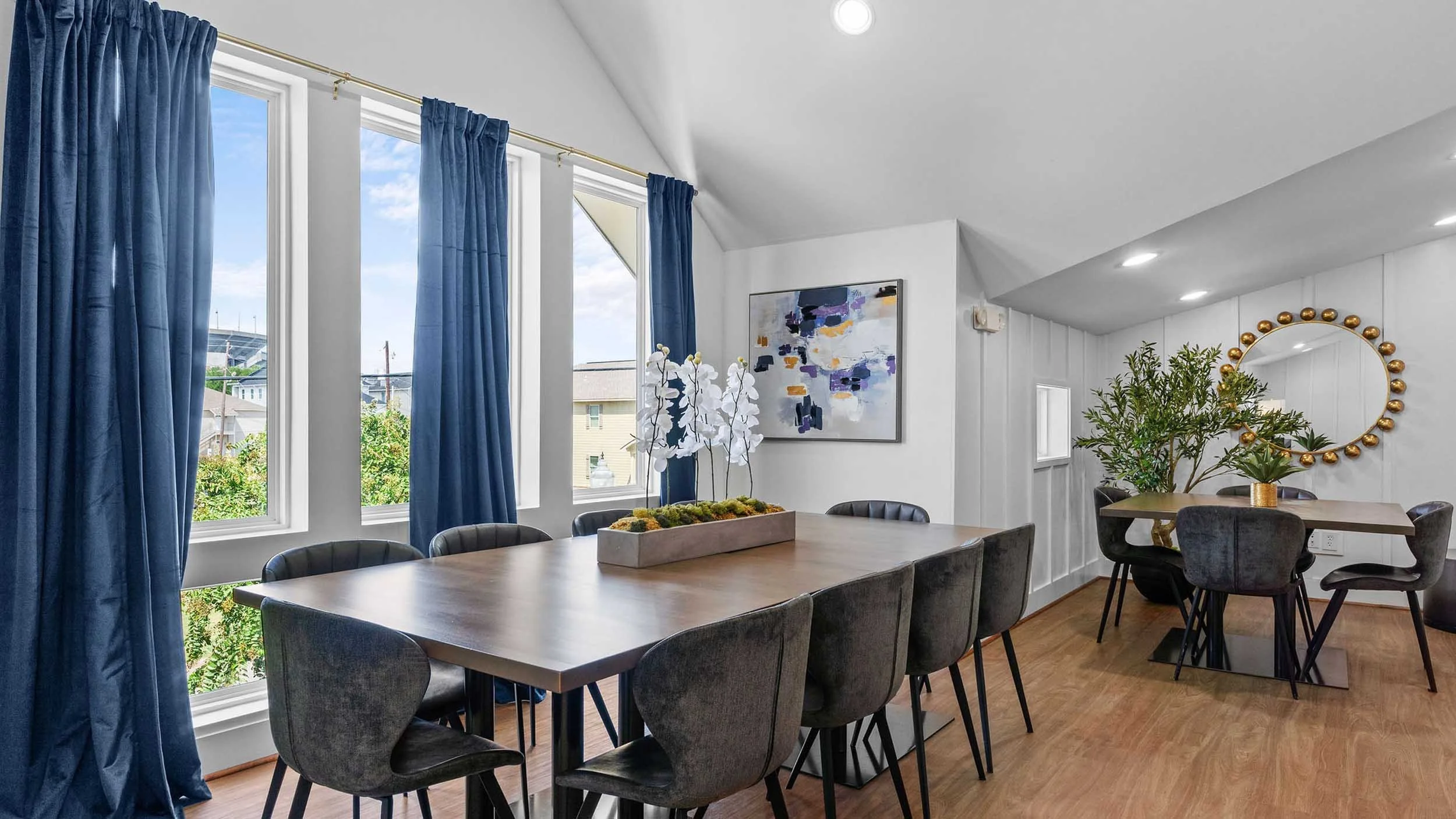 Study room with large windows and blue curtains, a long wooden table, black chairs, a modern abstract painting, potted plant, and a round mirror with gold embellishments on white paneled walls.
