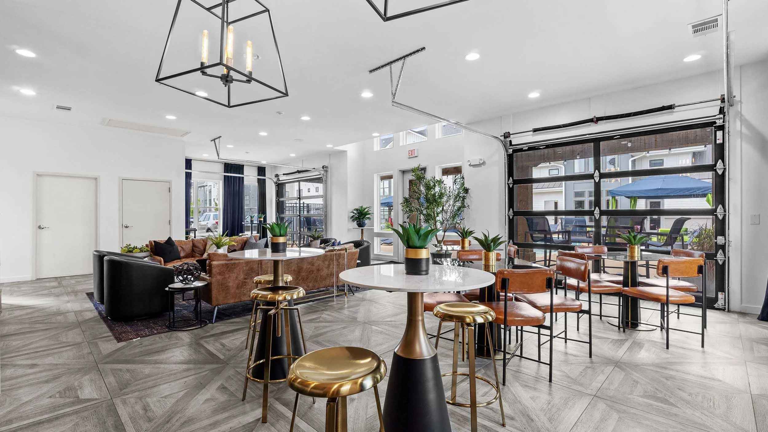 Bright, modern lounge area with various seating options including leather chairs, a sofa, and bar stools. There are multiple large potted plants, large windows, and skylights providing natural light. The space features track lighting, contemporary chandeliers, and a garage-style glass door leading outside.